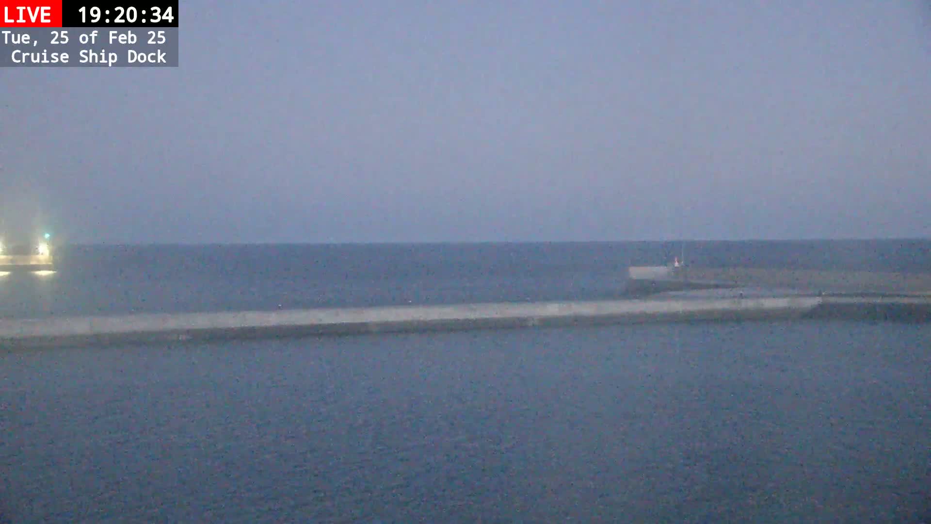 A long, low concrete wall stretches across a calm, dark-blue body of water under a dusky sky.