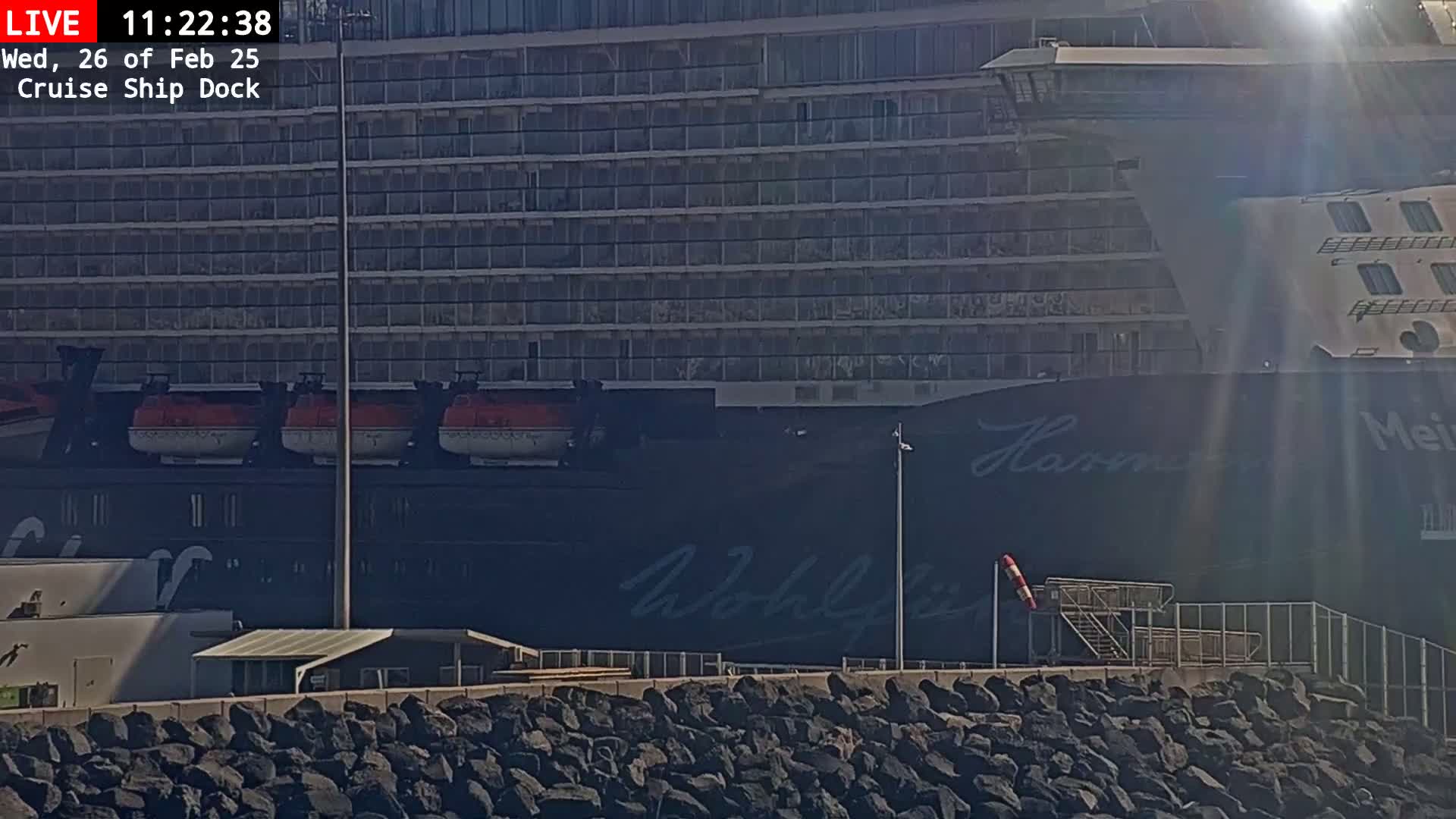 A large cruise ship is docked, partially obscured by bright sunlight, with a rocky embankment in the foreground.