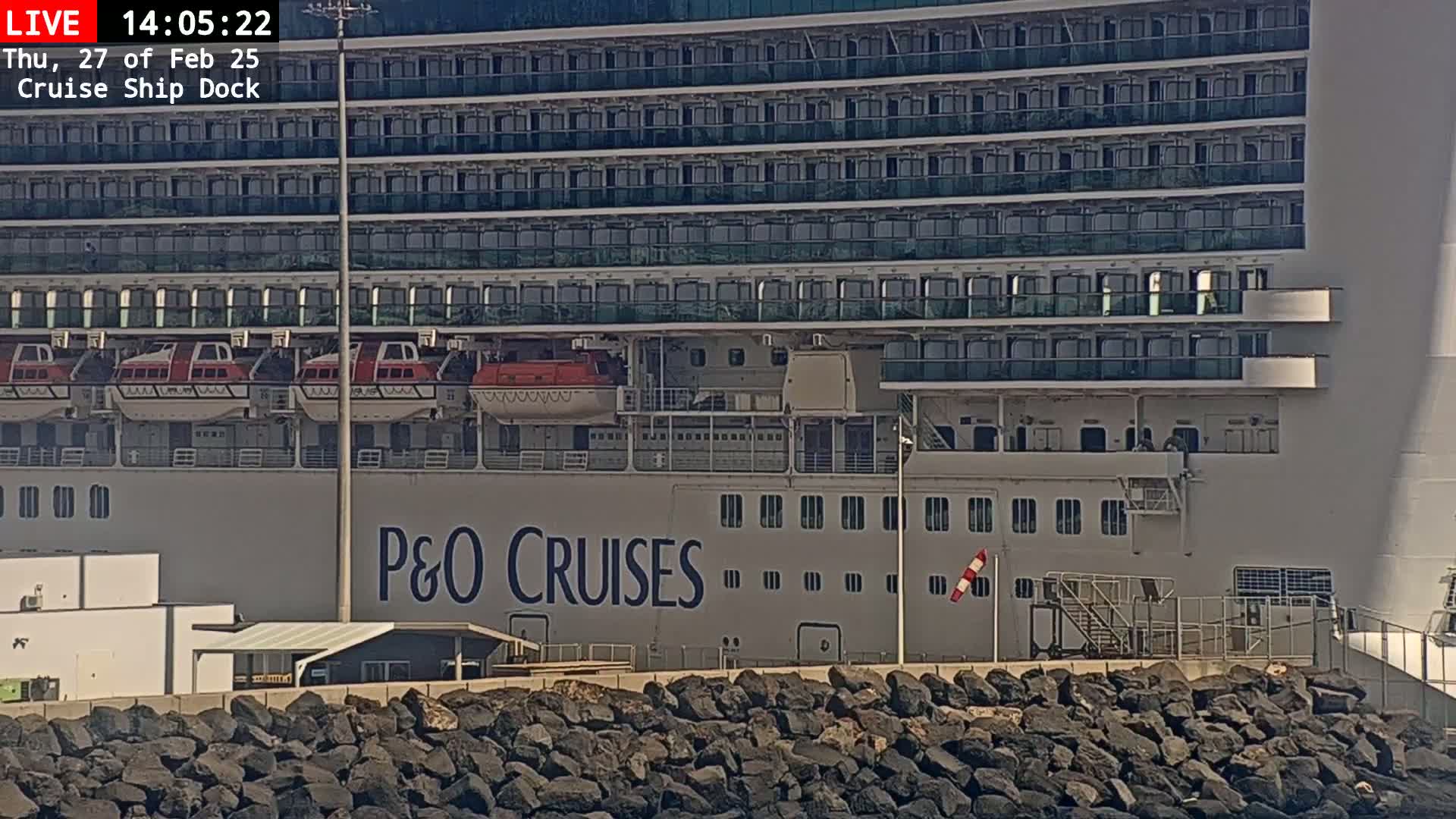 A large cruise ship, docked at a port on a sunny day, is seen from the side, showcasing its numerous decks and lifeboats.