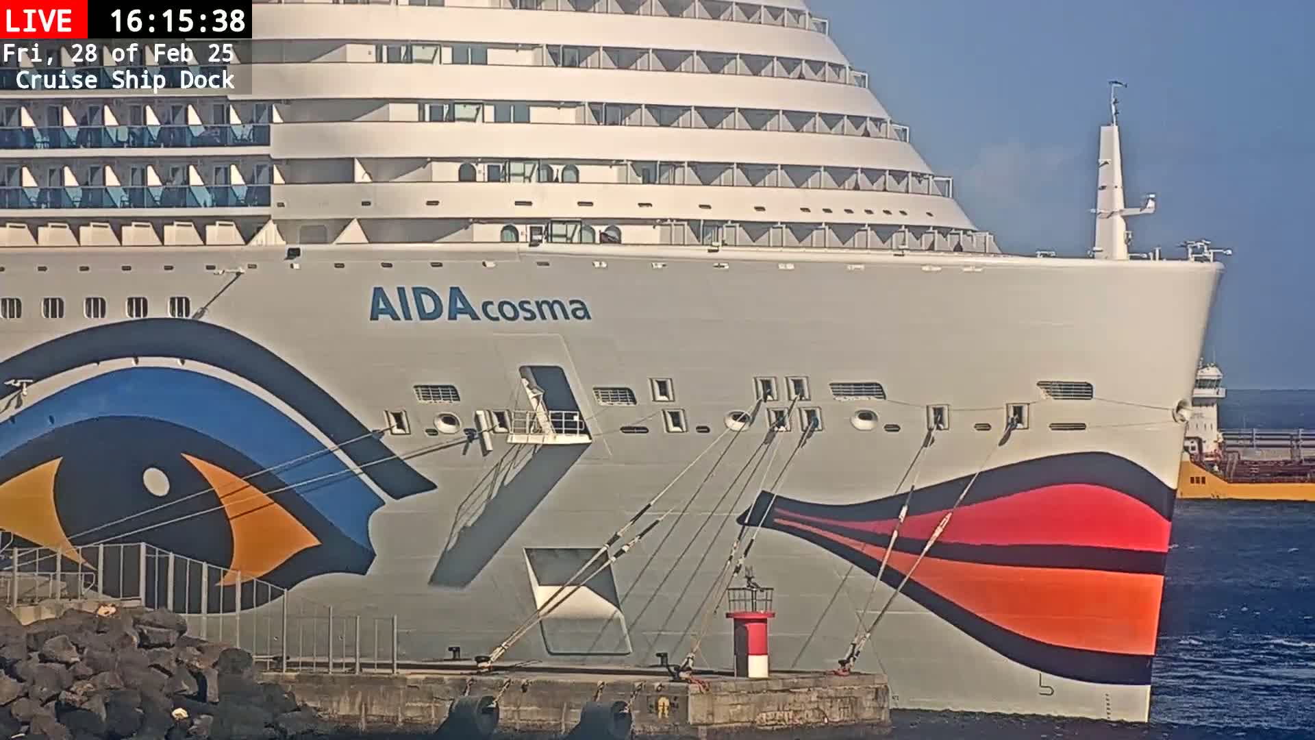 The bow of a large cruise ship with a stylized eye painted on its hull is docked, under sunny skies.