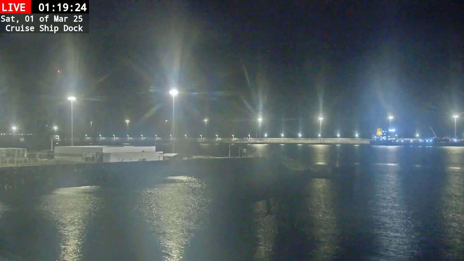 A nighttime view of a cruise ship docked at a pier, brightly lit by numerous lights, with calm water reflecting the lights.