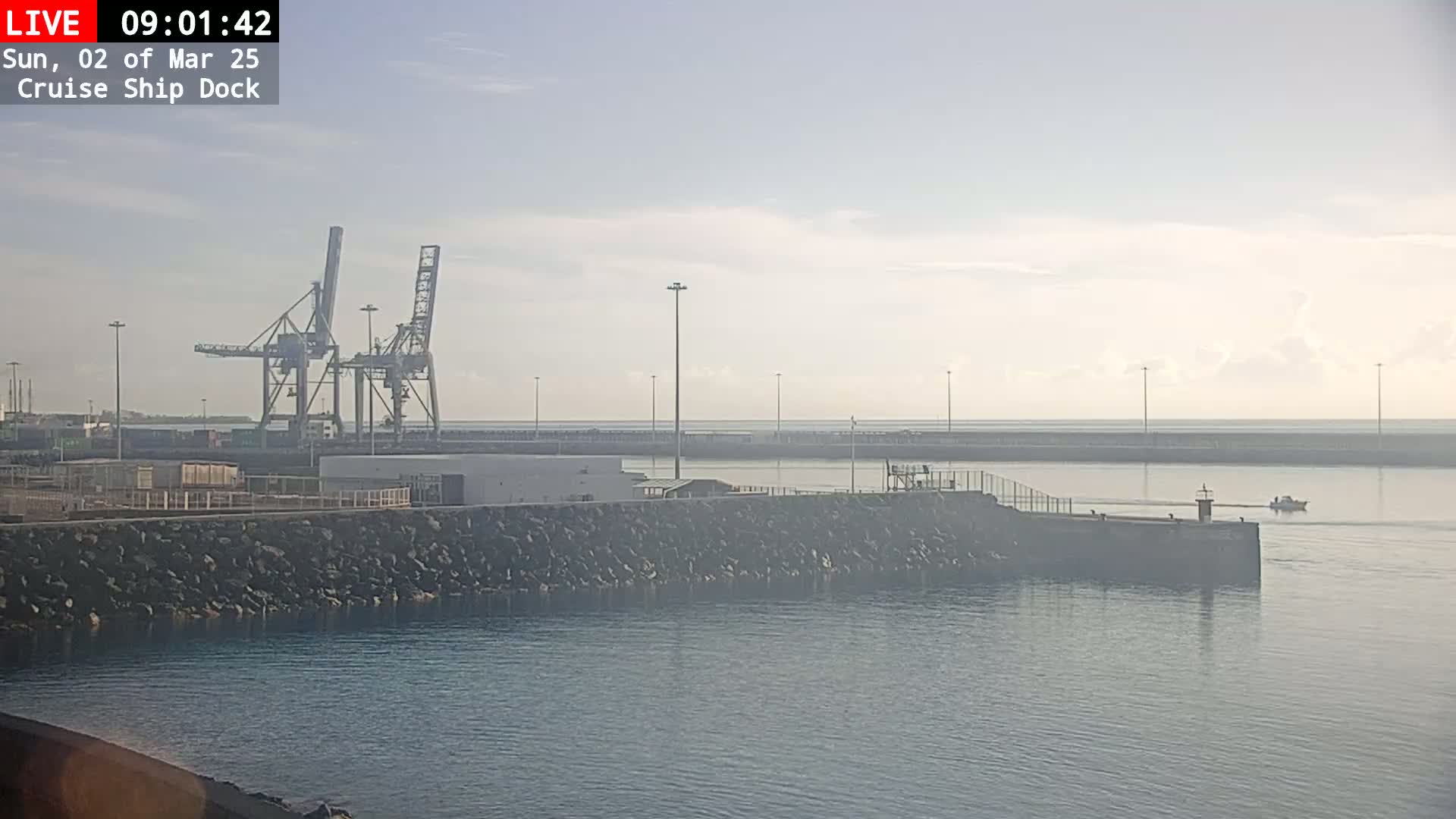 A calm, hazy harbor scene shows a cruise ship dock with cargo cranes, a rocky seawall, and a small boat in the distance.