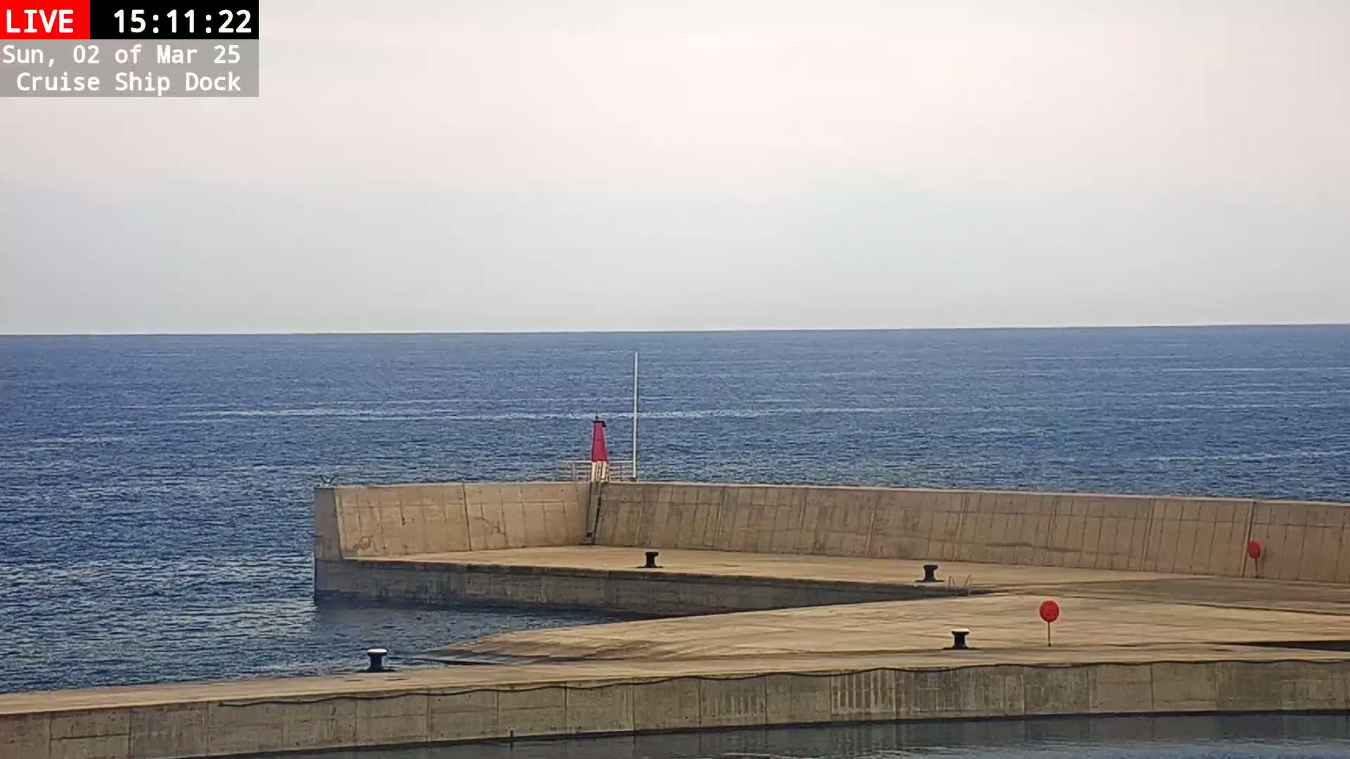 A calm, light-grey-skied day shows a concrete cruise ship dock extending into a dark blue sea, featuring a small red lighthouse.