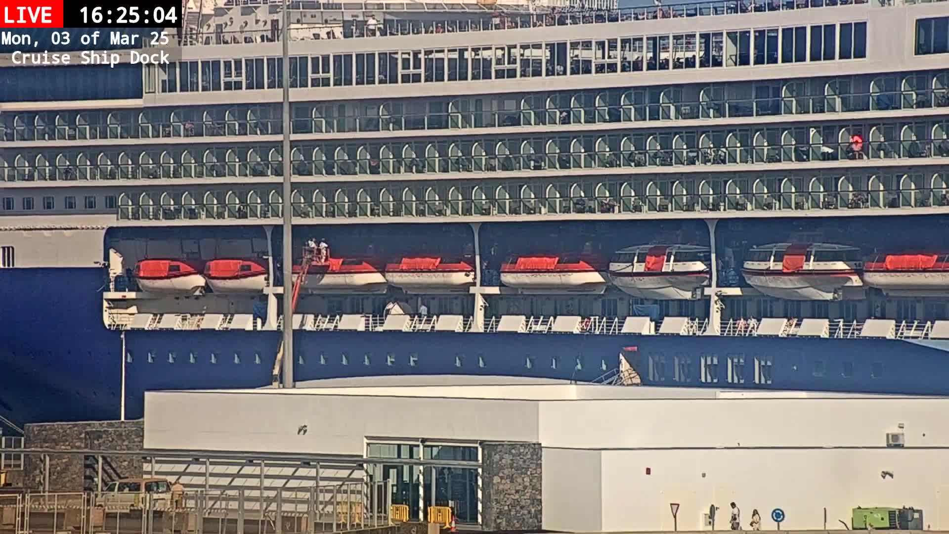 A large cruise ship is docked at a terminal on a sunny day, with several lifeboats visible on its side.