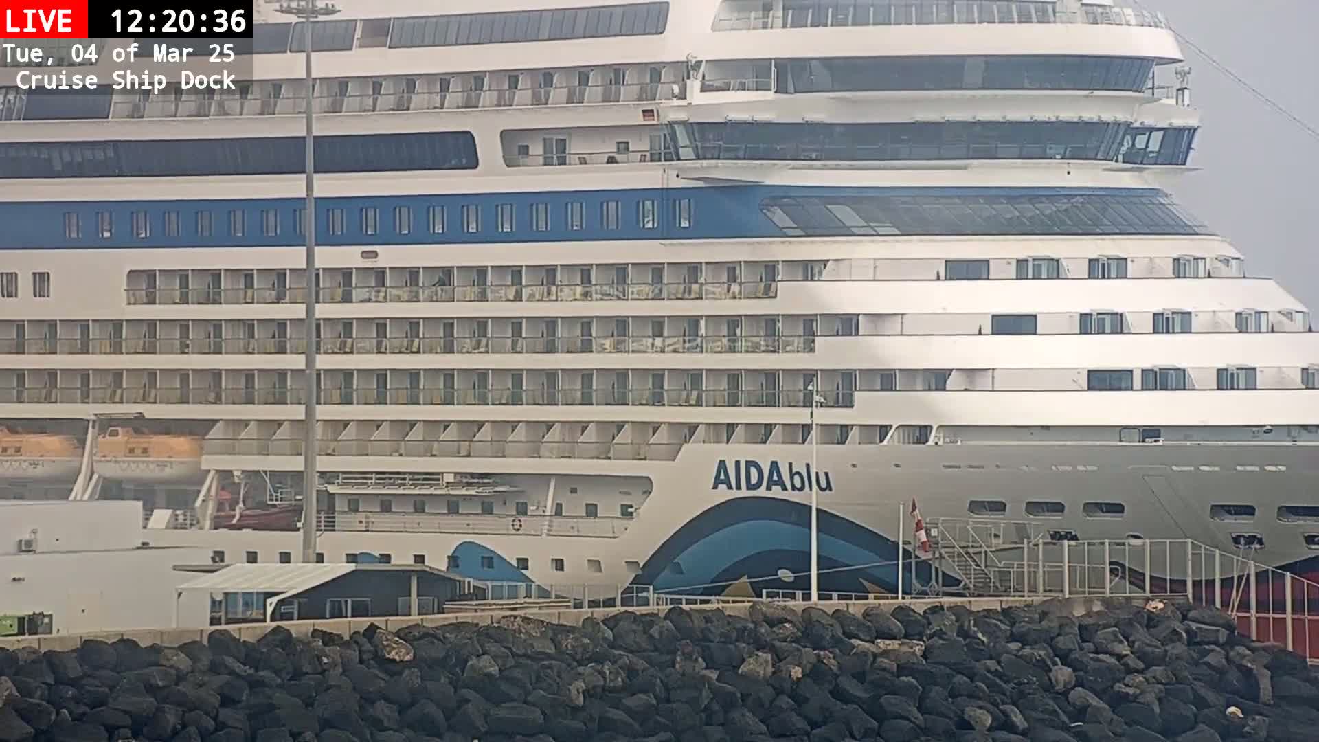 A large cruise ship is docked at a port on an overcast day, behind a dark rock wall.