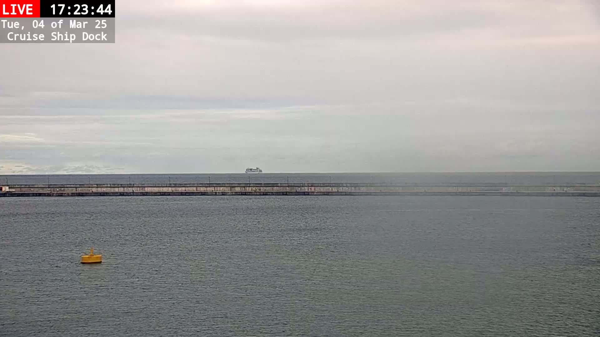 A cruise ship is visible on the horizon beyond a long, low sea wall in overcast conditions.
