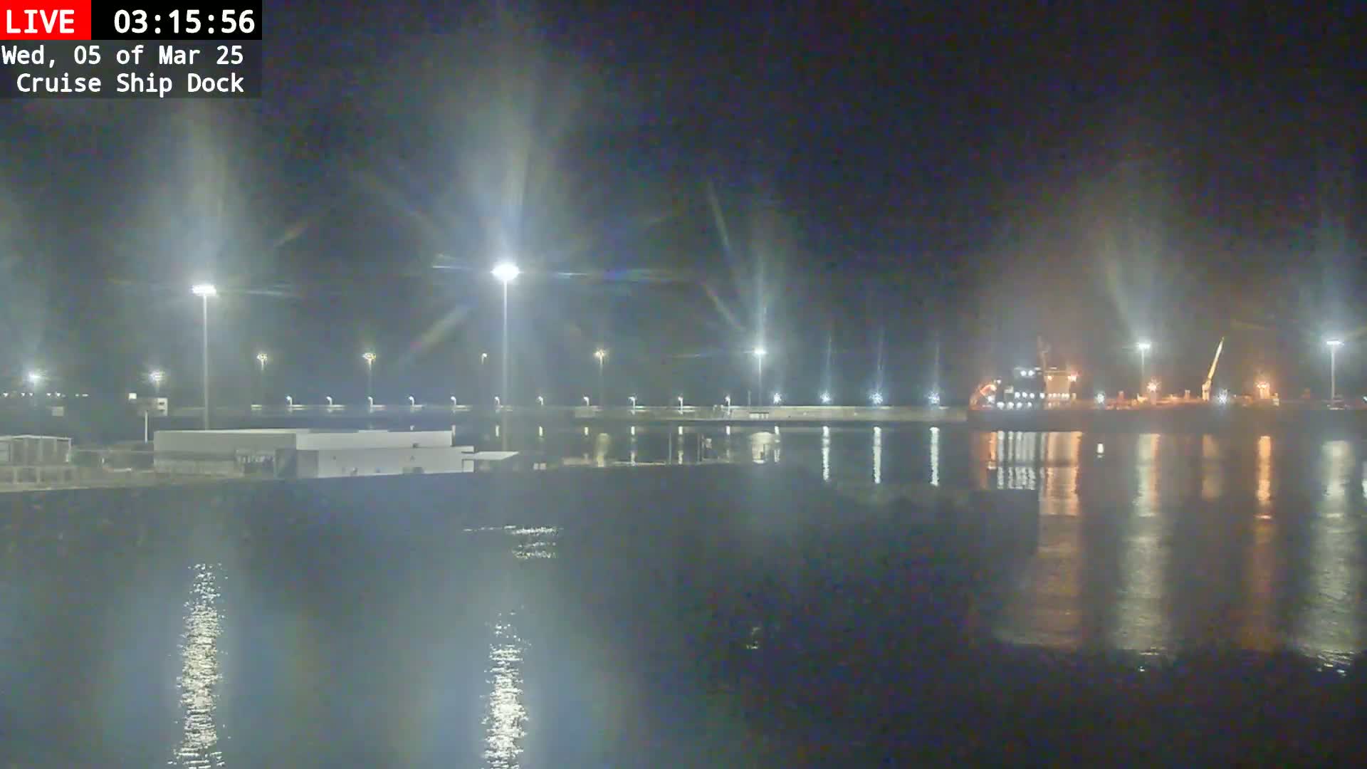 A nighttime view of a cruise ship dock, illuminated by bright lights, with a large ship moored nearby and calm water reflecting the lights.