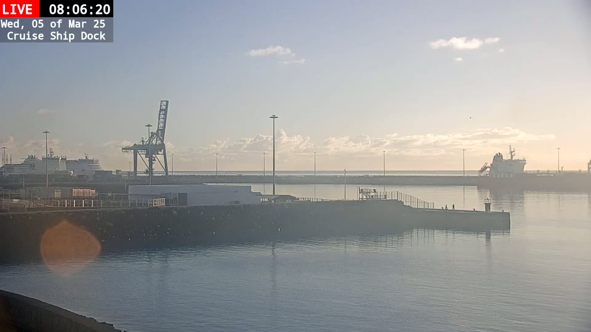 A calm harbor at sunrise shows several ships docked near a large crane, with a hazy atmosphere.