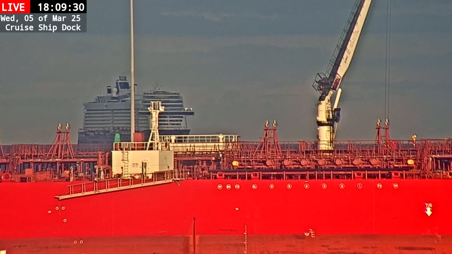 A large red tanker ship is docked next to a cruise ship under an overcast sky.