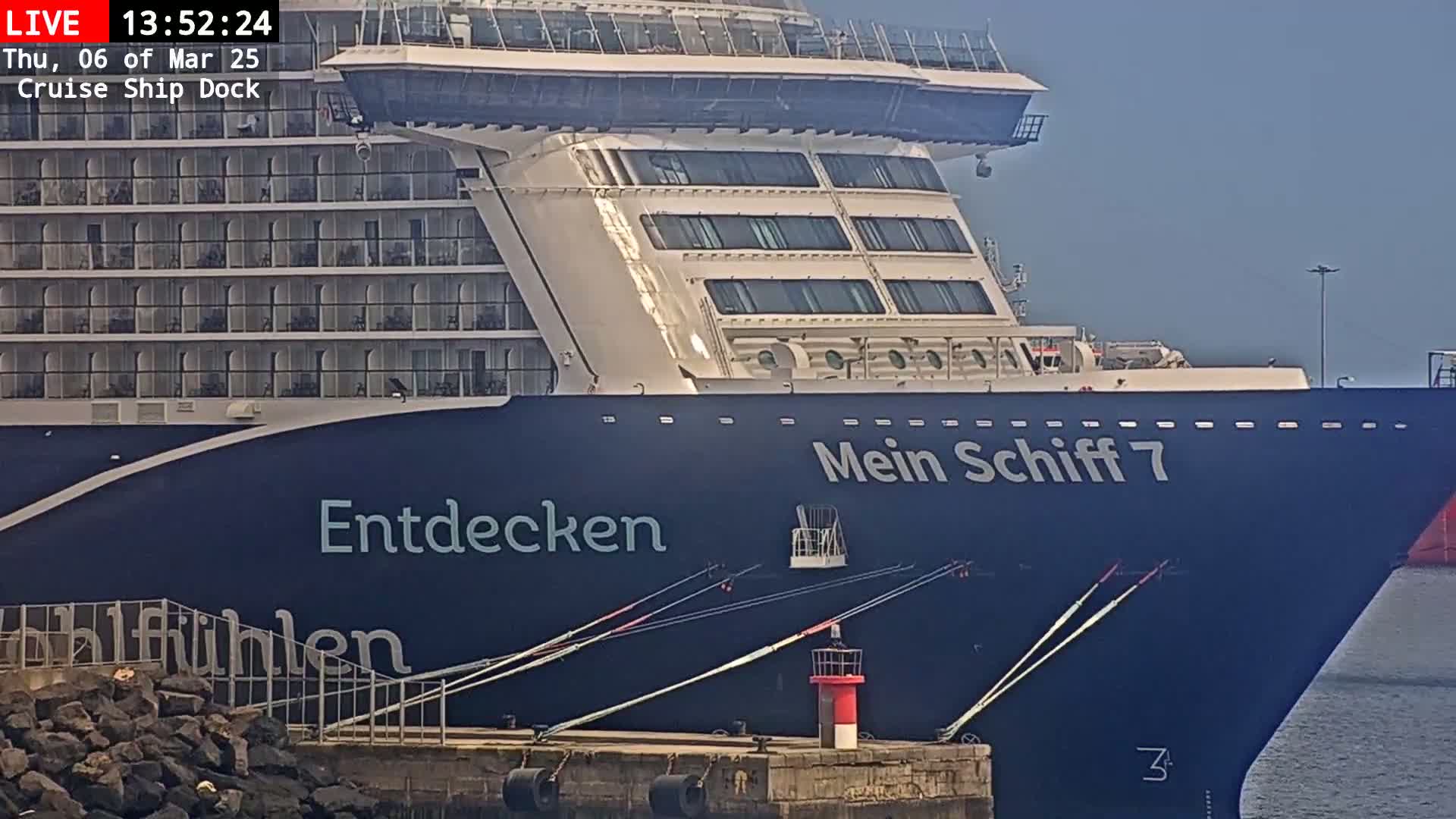 A large cruise ship, docked and secured by ropes, is shown on a partly cloudy day.