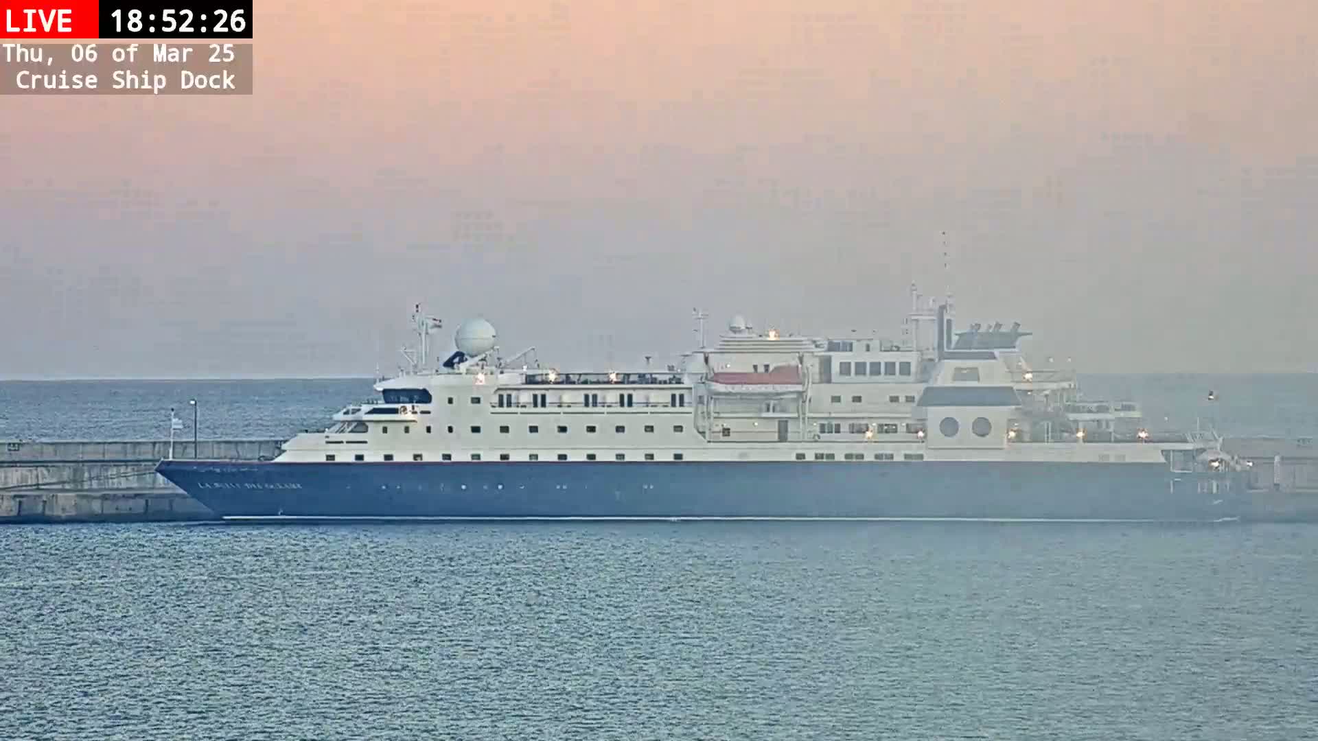A large cruise ship is docked at a pier on a calm, hazy evening.