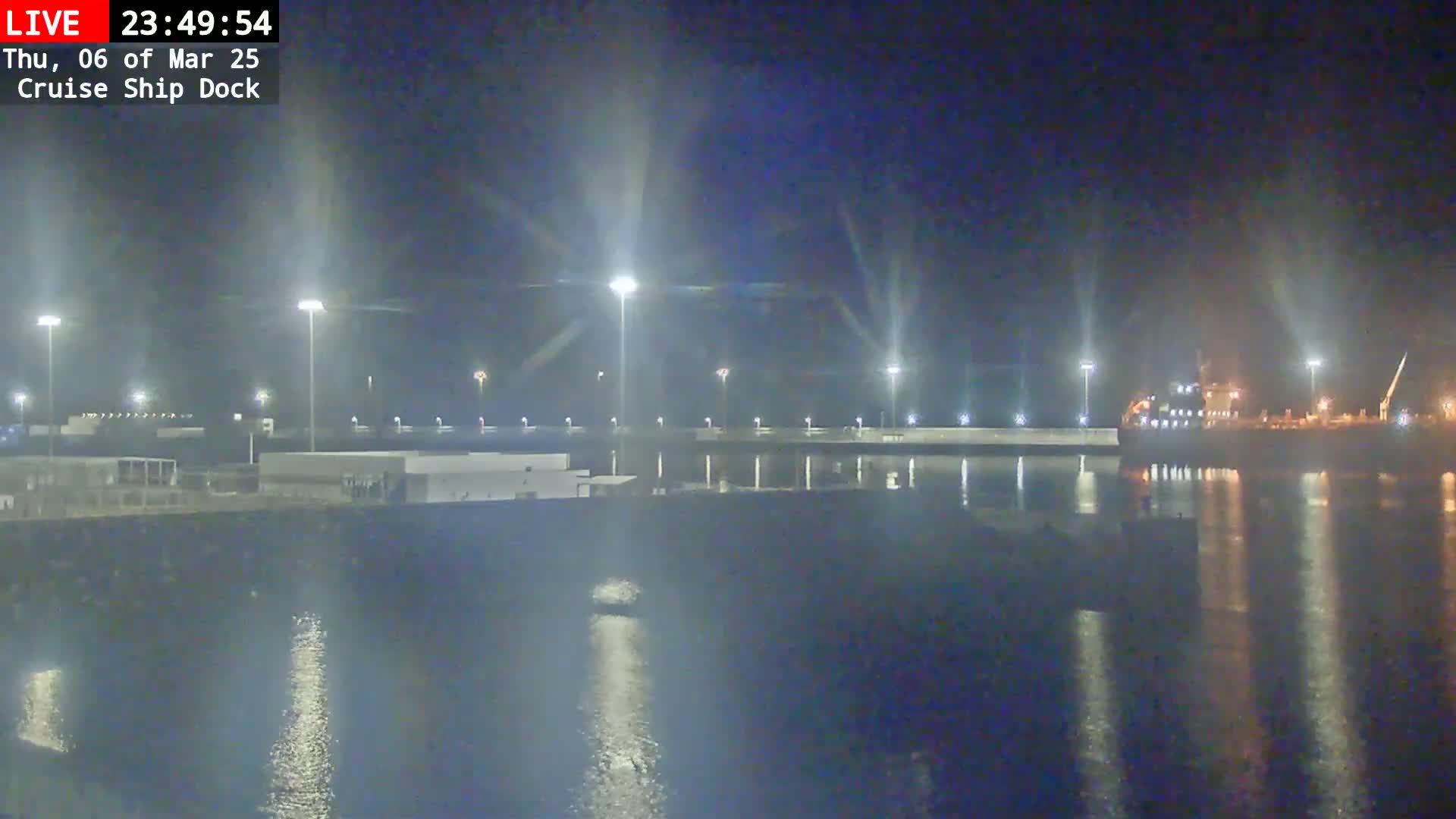 A nighttime view of a cruise ship docked at a port, illuminated by bright lights reflecting on calm water.