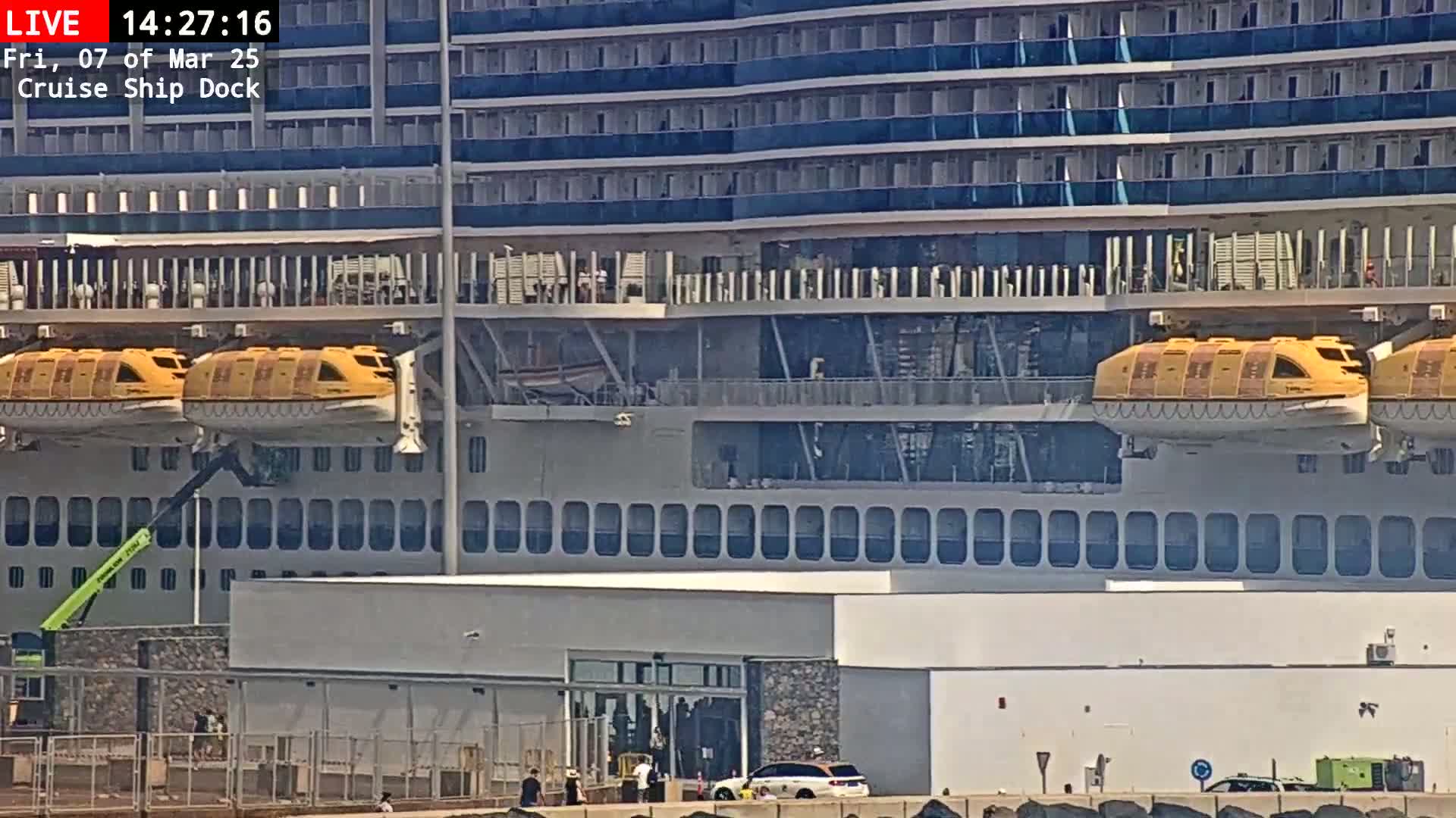 A large cruise ship is docked, with two yellow lifeboats visible, under sunny conditions.