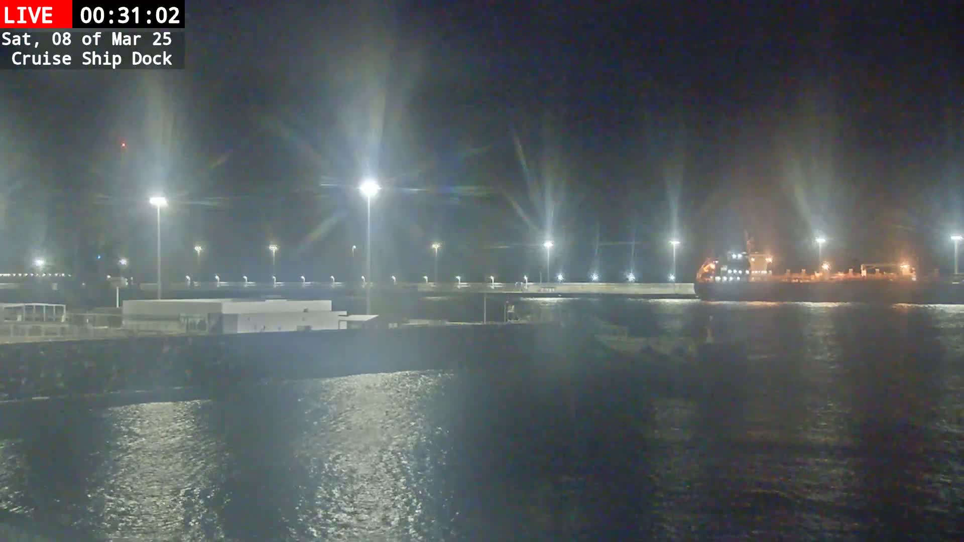 A large ship is docked at a brightly lit cruise ship terminal at night, with calm water visible.