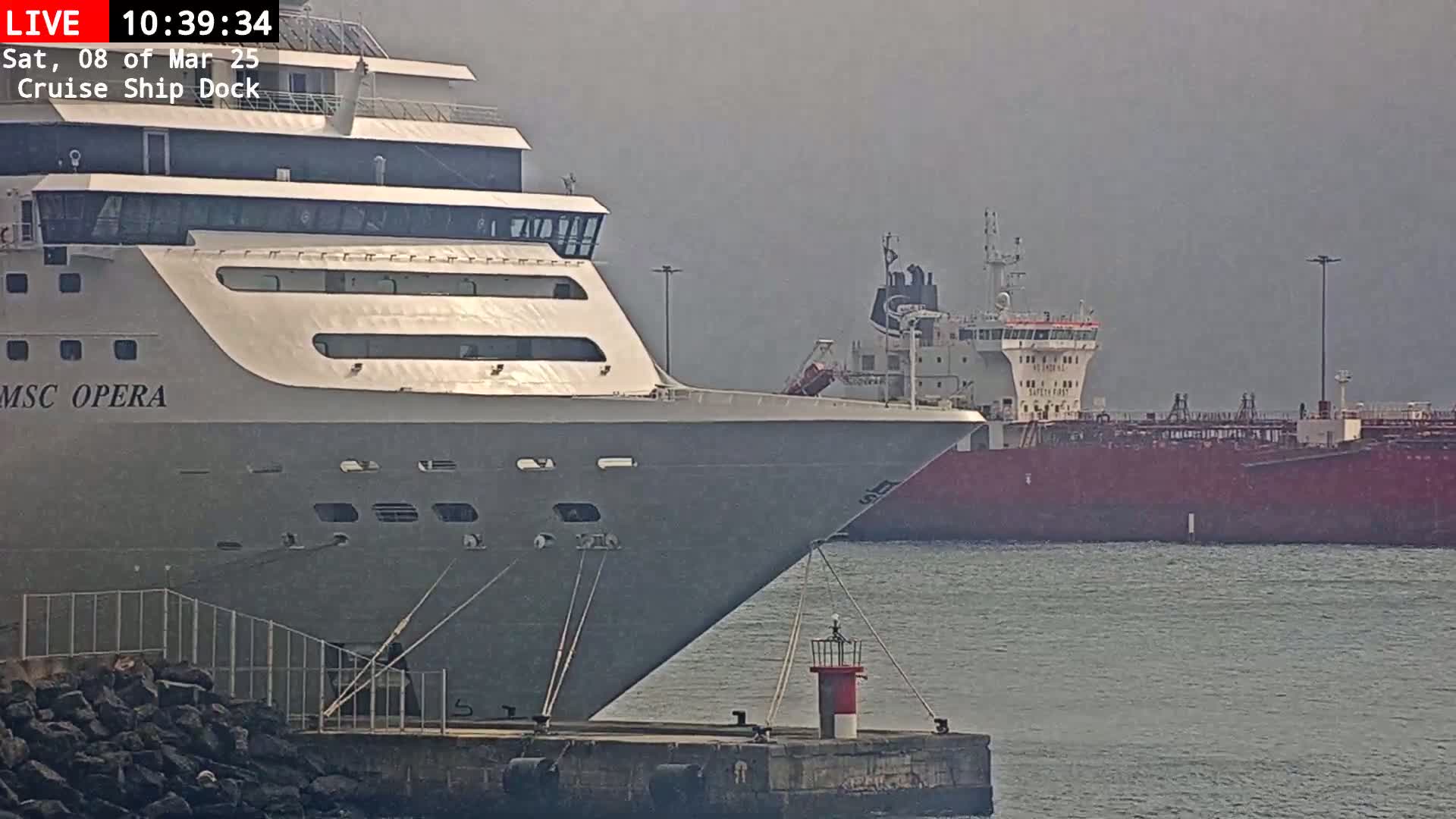 A large cruise ship is docked, with a smaller tanker visible in the background on a hazy day.