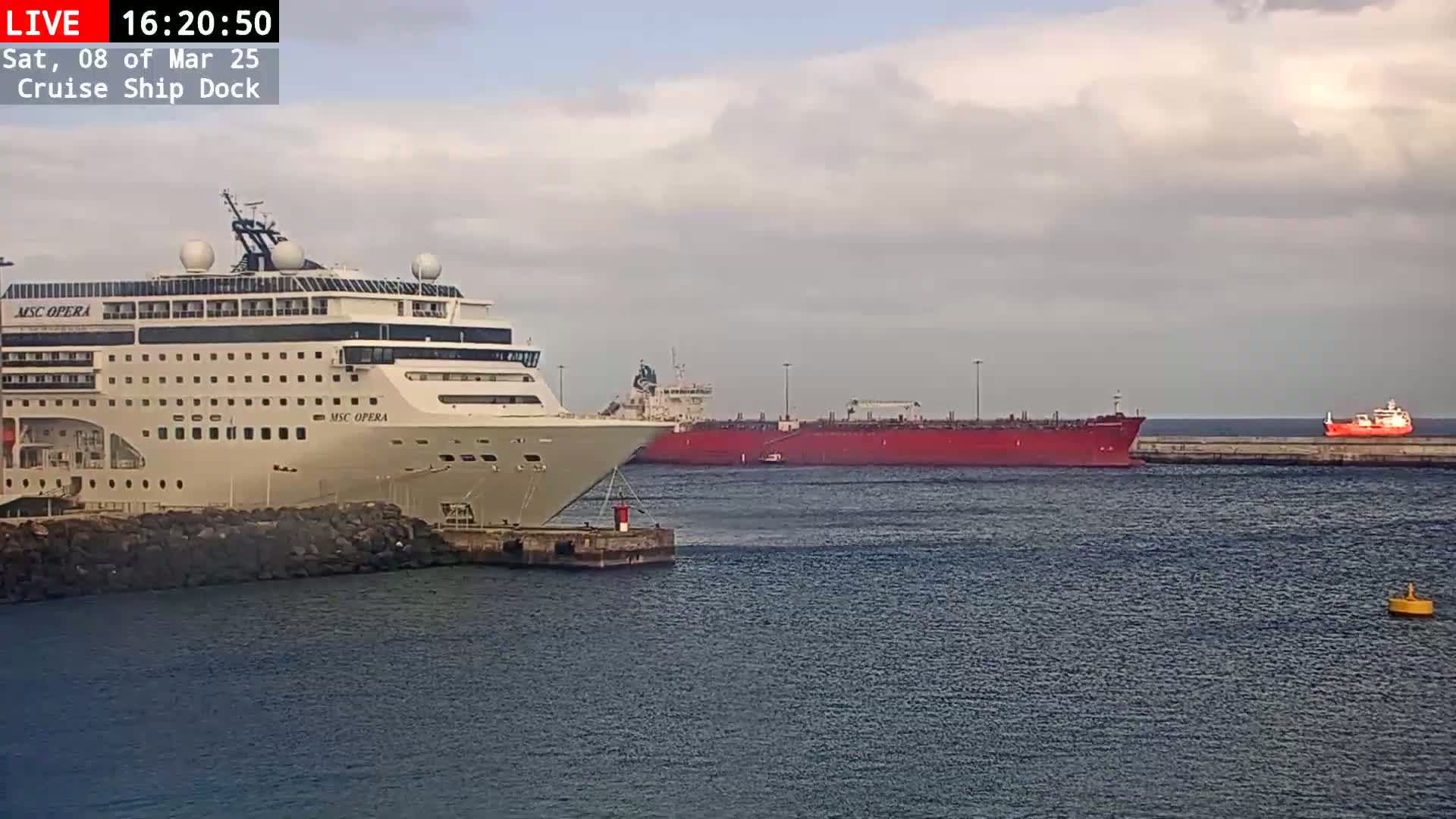 A large cruise ship is docked next to a red tanker ship in a calm harbor under a mostly cloudy sky.
