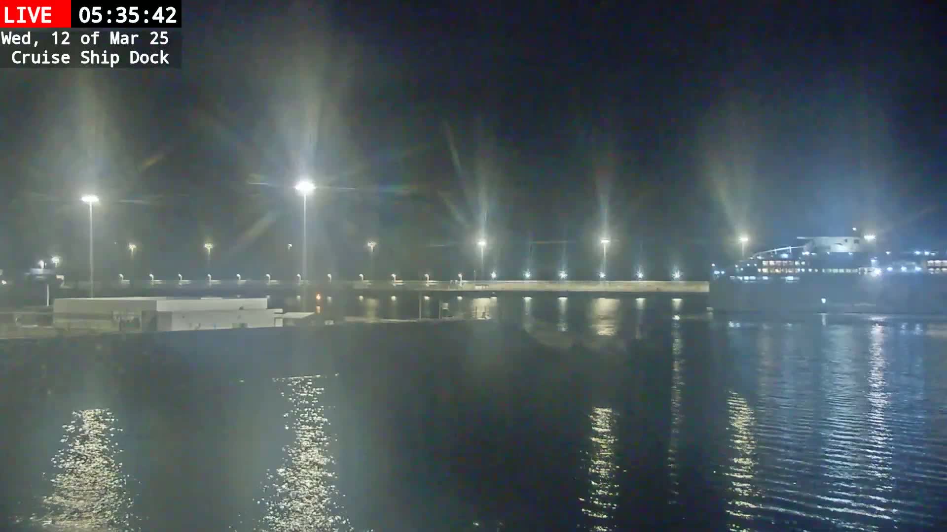 A nighttime view of a cruise ship docked at a pier, illuminated by bright lights reflecting on calm water.