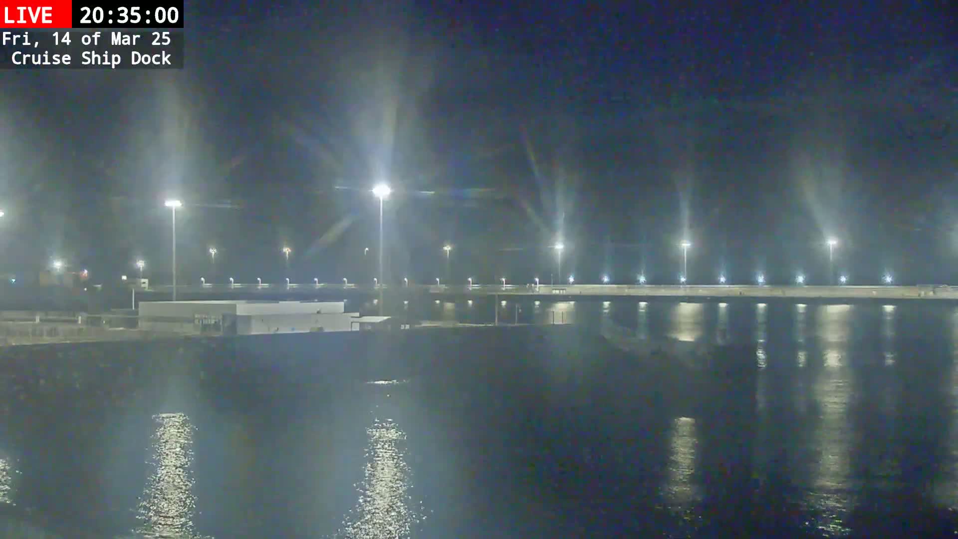 A nighttime view of a brightly lit cruise ship dock reflecting in calm water under a dark sky.