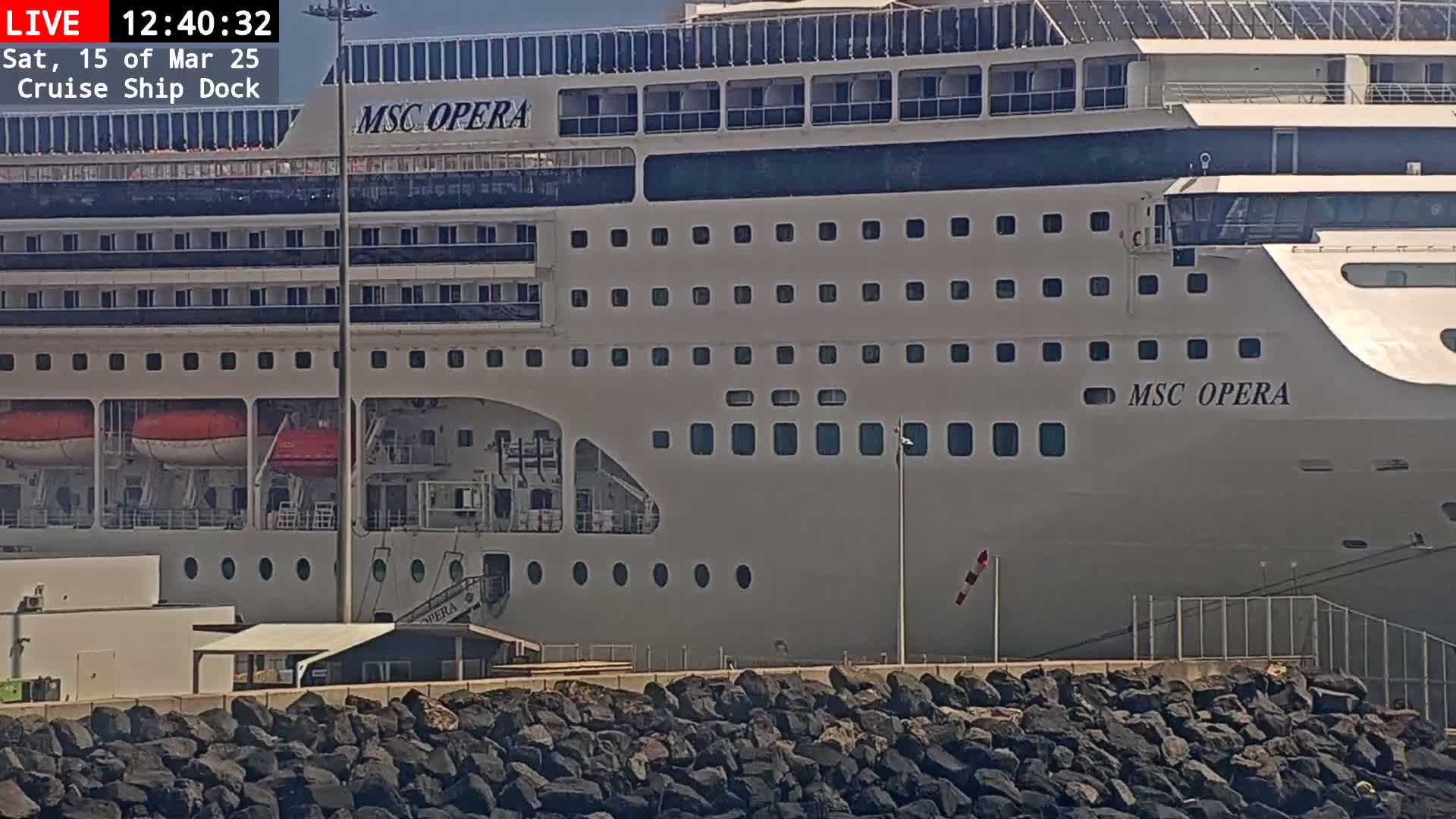 A large cruise ship, the MSC Opera, is docked at a port on a sunny day, with a rocky embankment in the foreground.