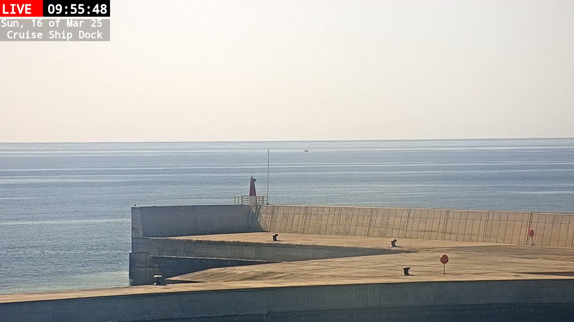 A concrete cruise ship dock extends into a calm, light-blue sea under a hazy sky.