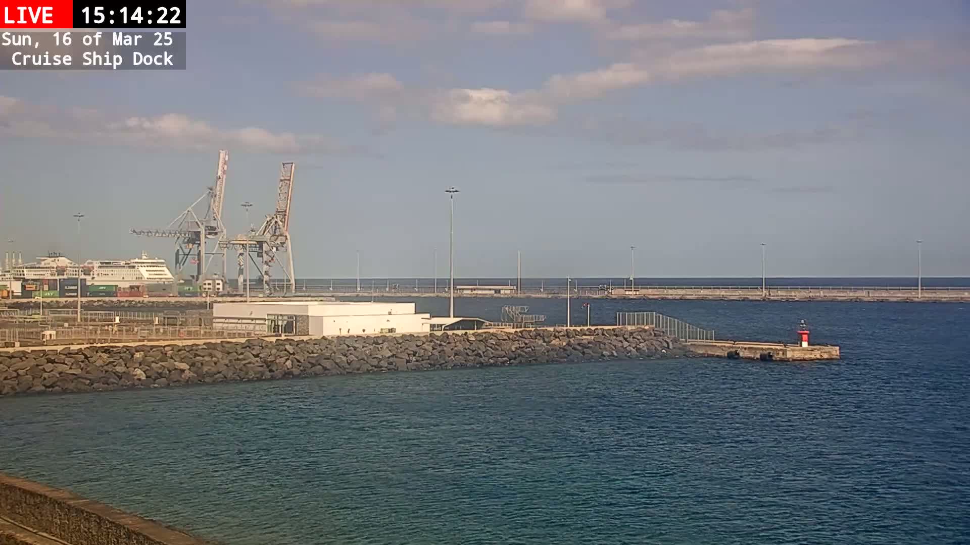 A cruise ship is docked at a port under a partly cloudy sky, with large cranes visible in the background.