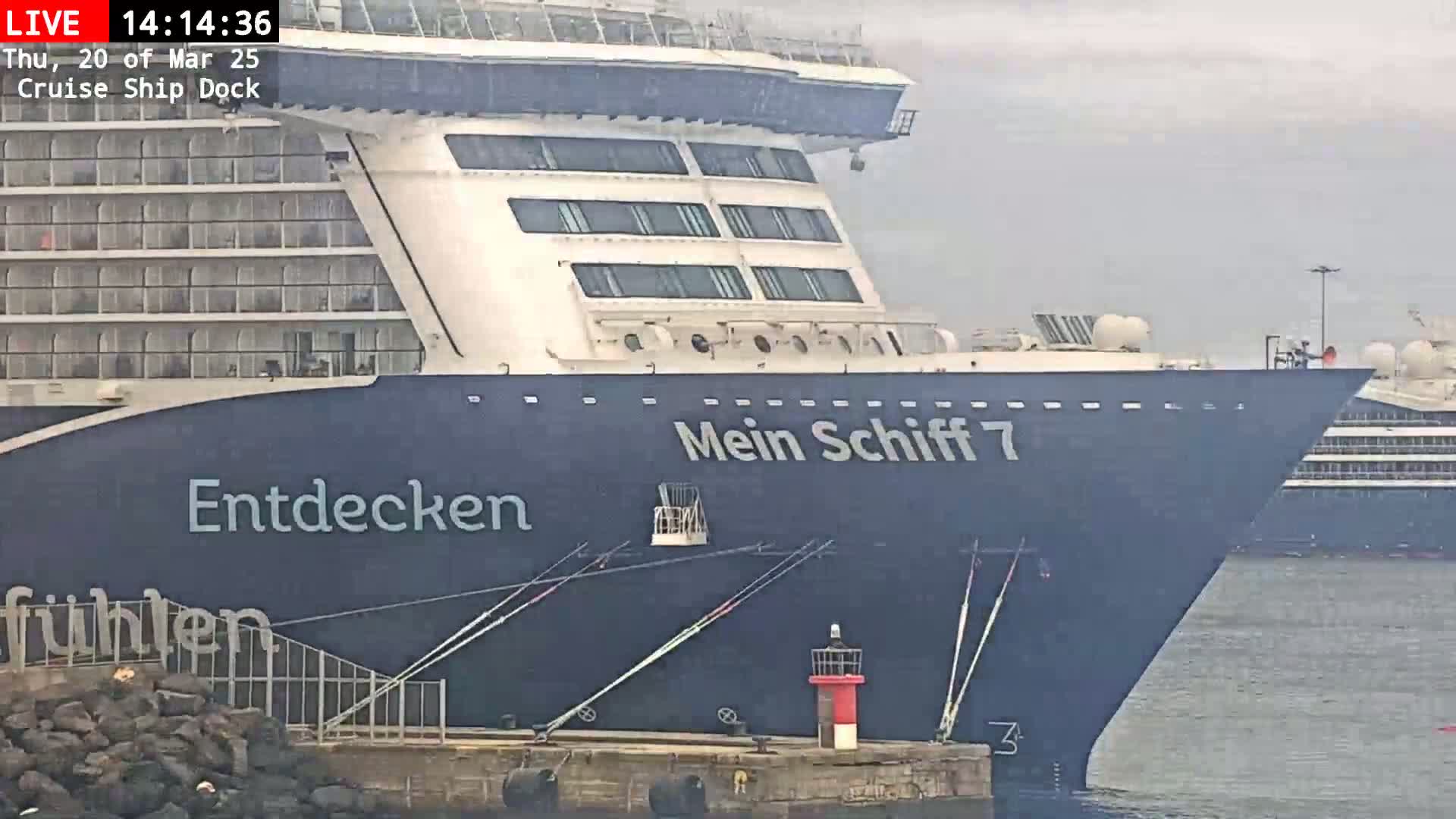 A large cruise ship, named "Mein Schiff 7," is docked, with overcast skies.