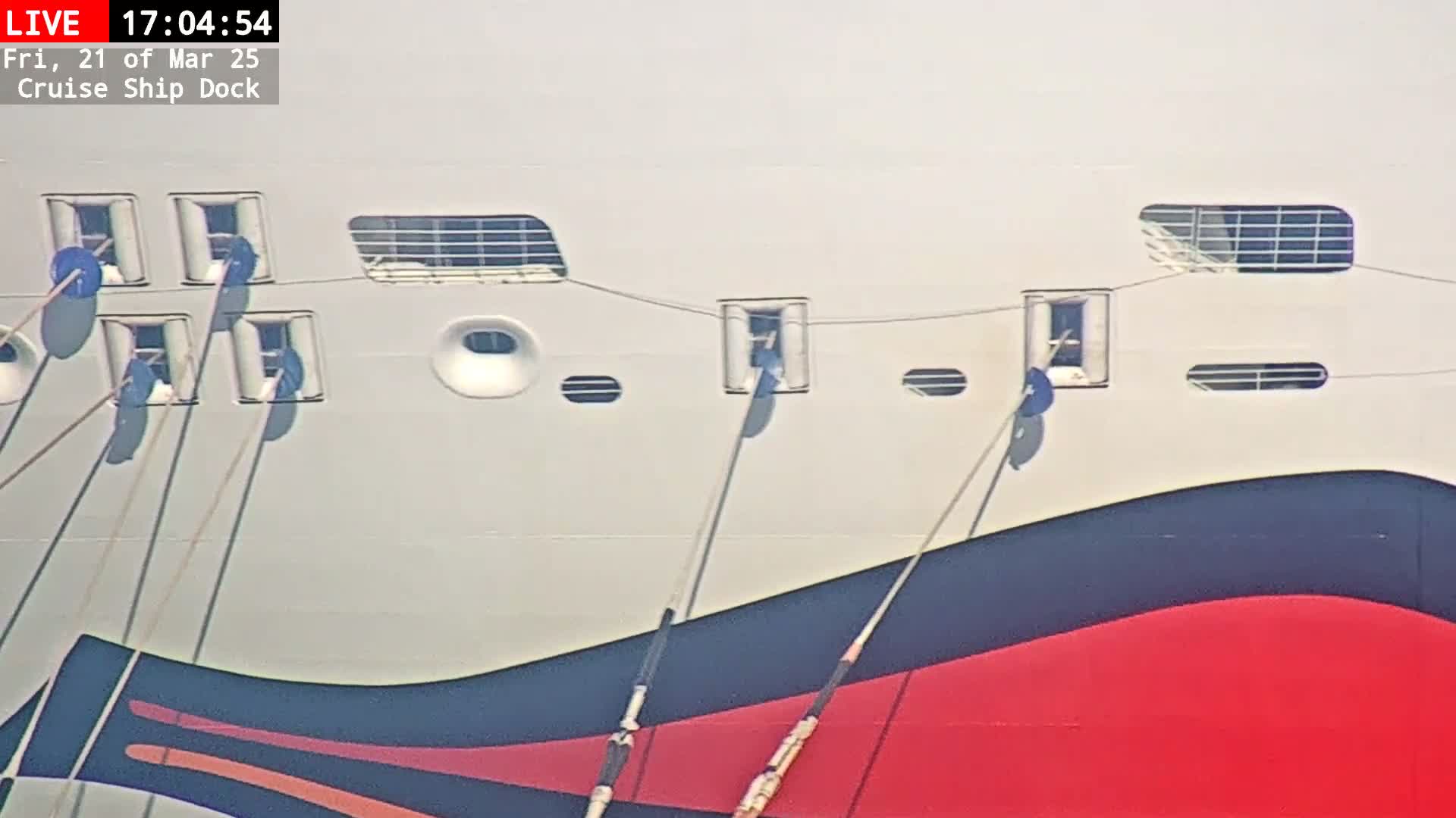 The lower portion of a large, predominantly white cruise ship's hull is shown, featuring several small windows and mooring lines against a bright, sunny sky.