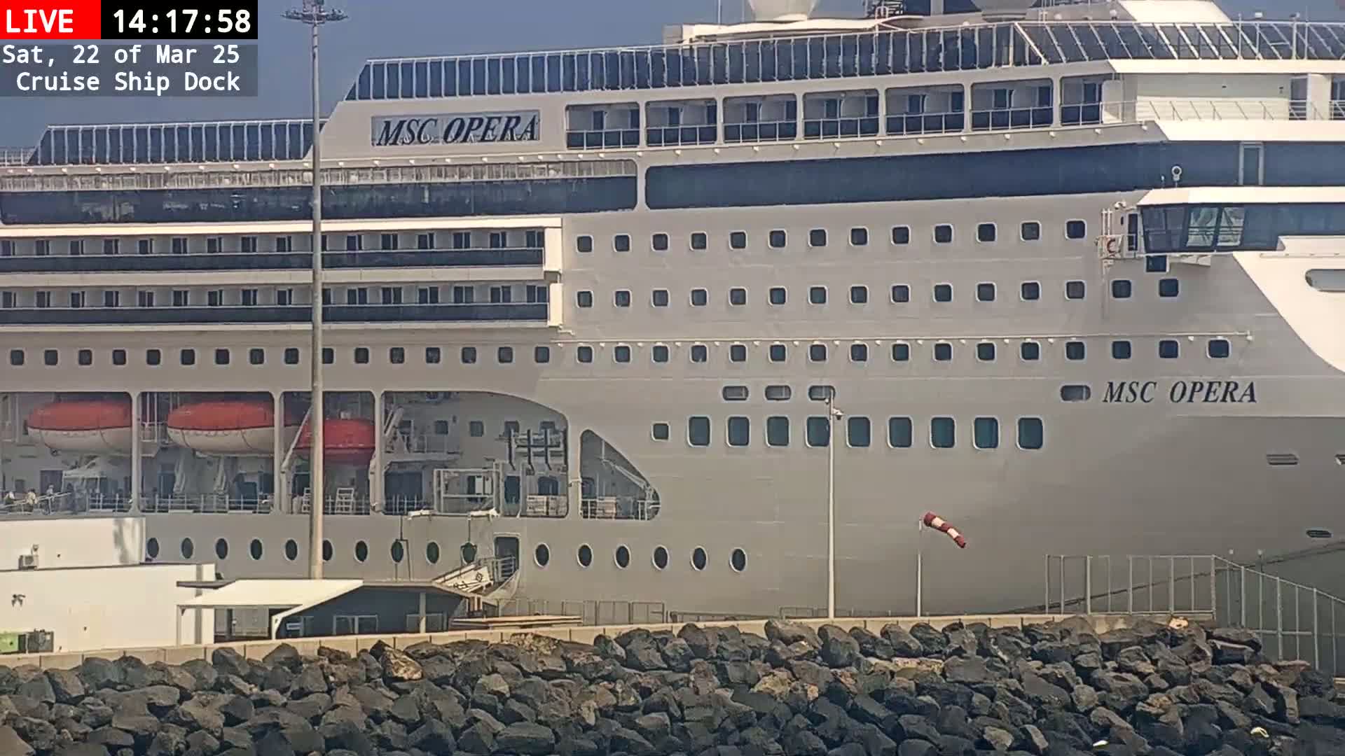 A large cruise ship is docked, with a mostly sunny sky and a windsock showing a slight breeze.