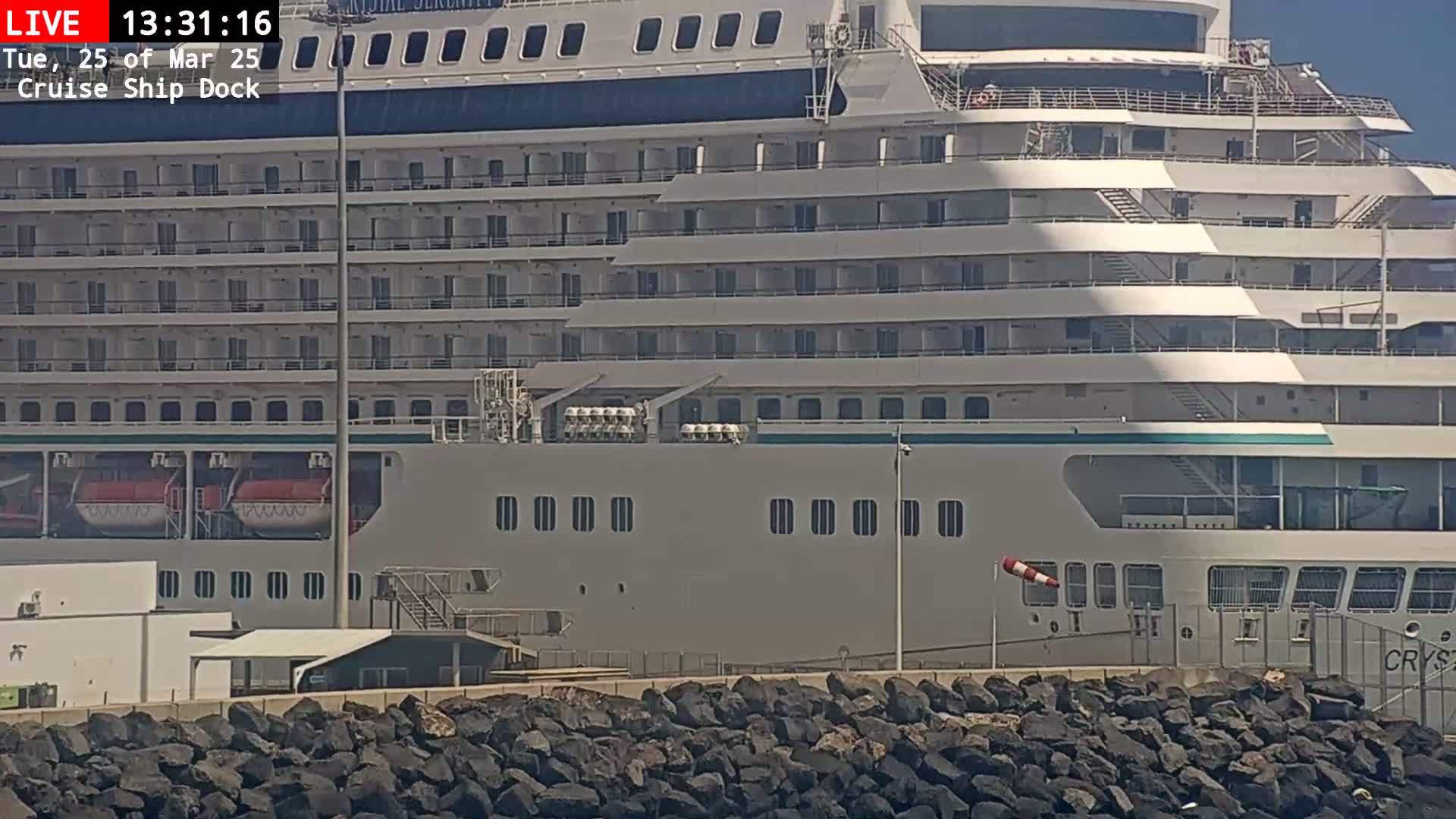 A large cruise ship is docked at a port on a sunny day, with a rocky embankment in the foreground.