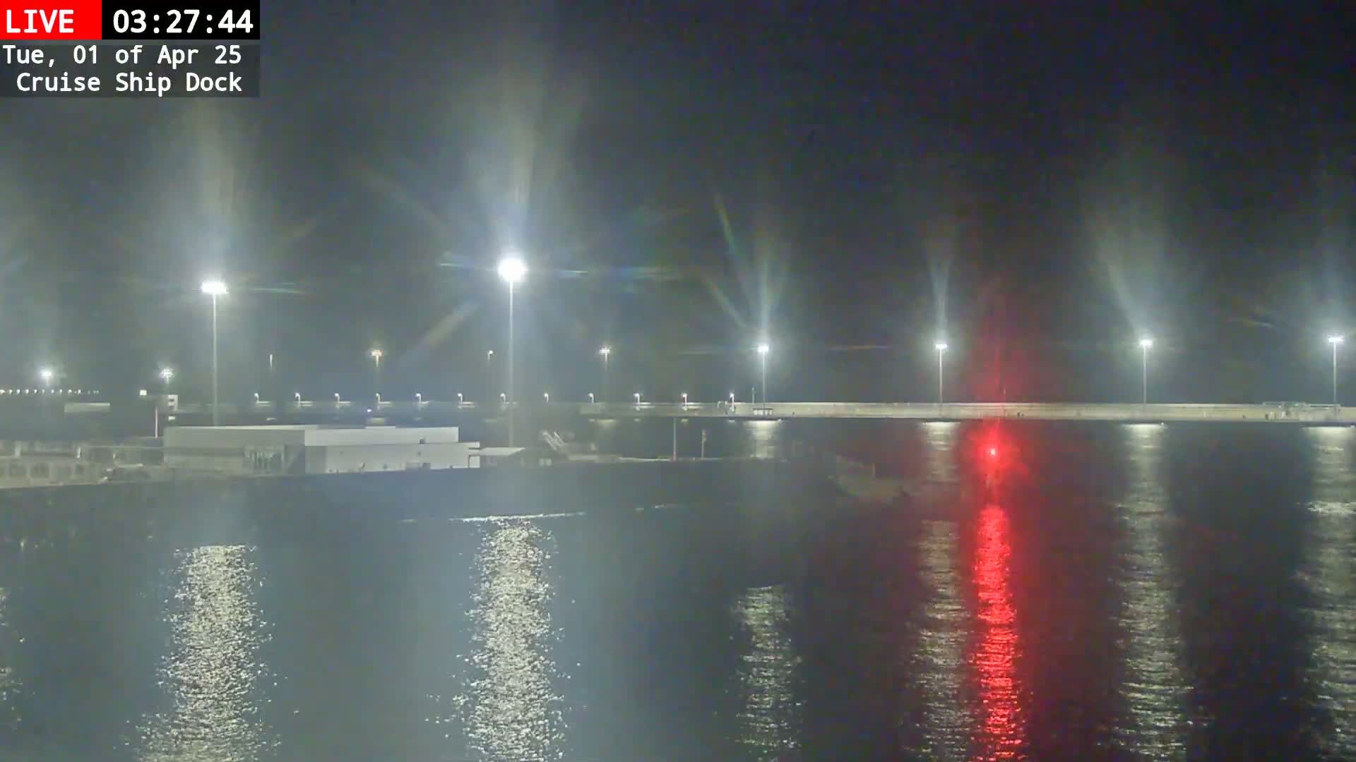 A nighttime view of a cruise ship dock illuminated by bright lights, reflecting on calm water, with a single red light visible on the water's surface.