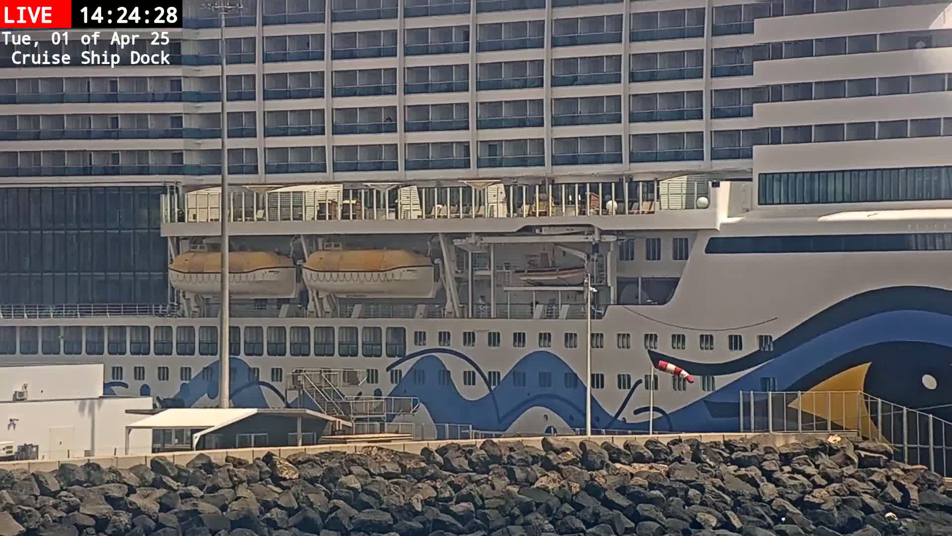 A large cruise ship is docked, with lifeboats visible, next to a rocky shore on a sunny day.
