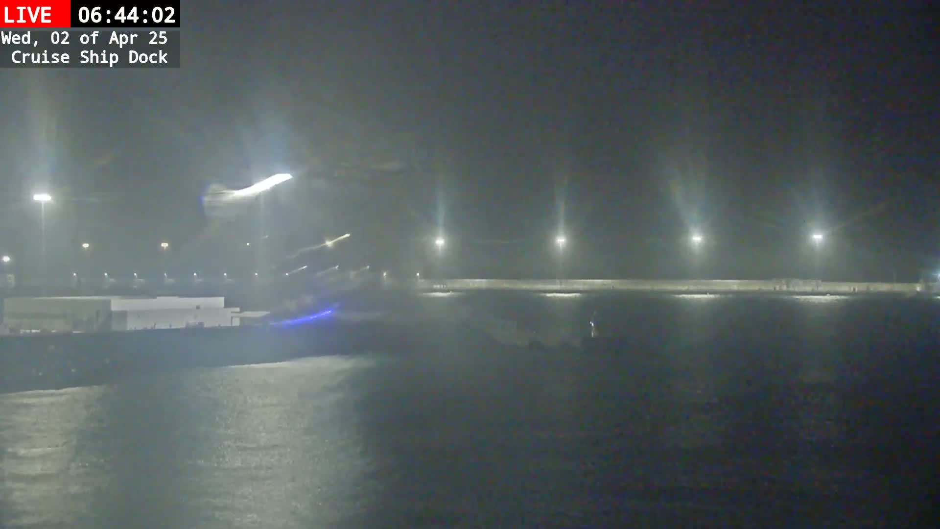 A nighttime, foggy view of a cruise ship dock with a large vessel partially visible and lights illuminating a wall along the water.