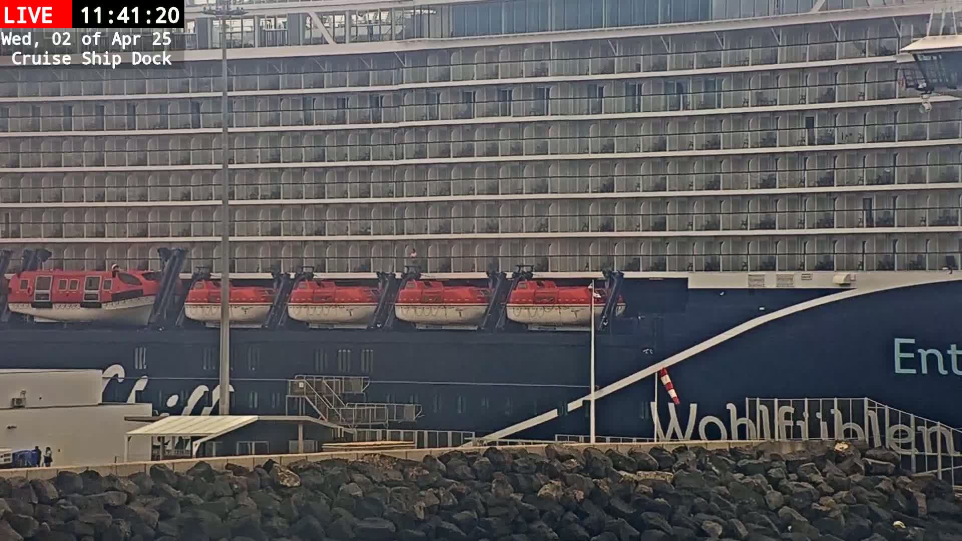 A large cruise ship is docked, its multiple lifeboats visible, next to a rocky shoreline on a cloudy day.