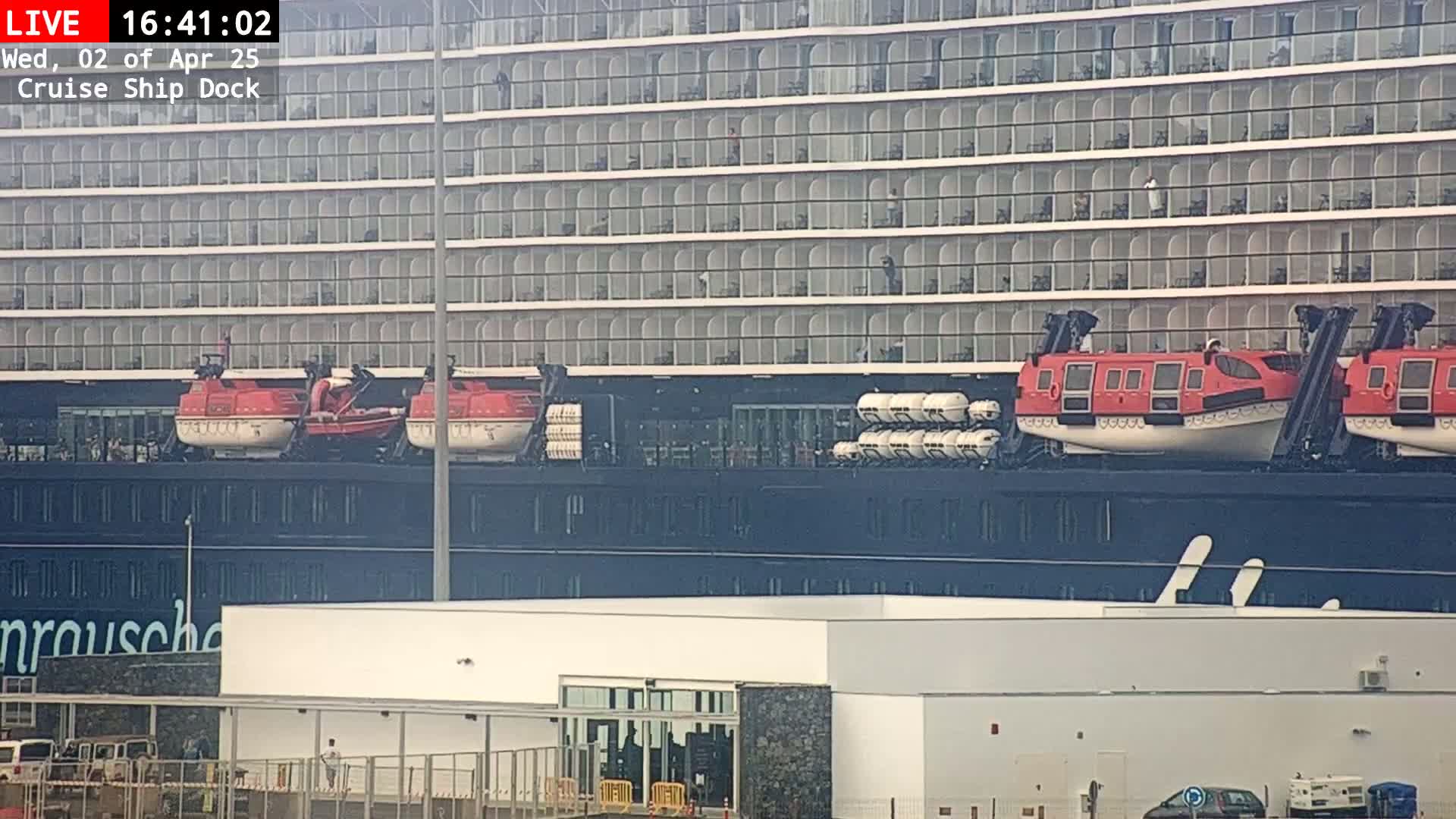 A large cruise ship is docked, with several lifeboats visible on deck, and the sky is overcast.