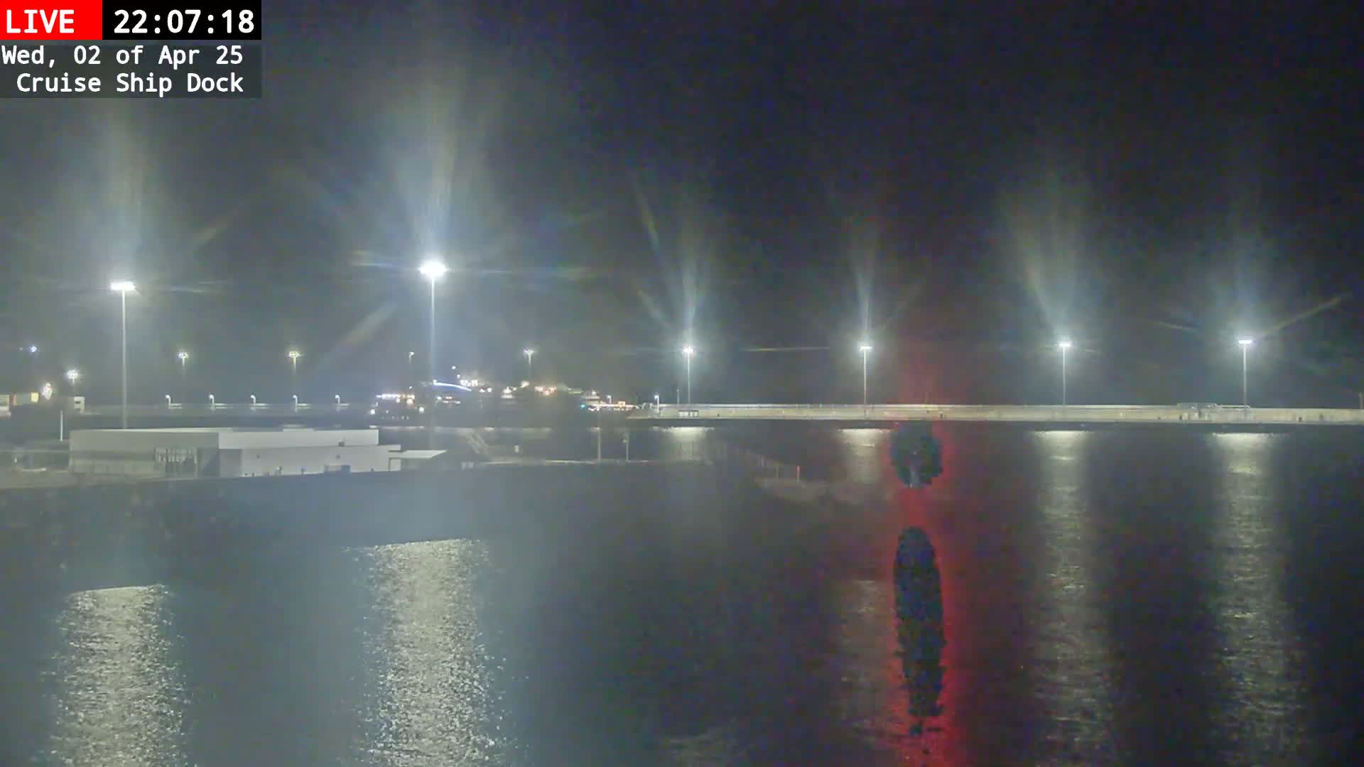 A nighttime view of a cruise ship dock with calm water reflecting bright lights, under a clear, dark sky.