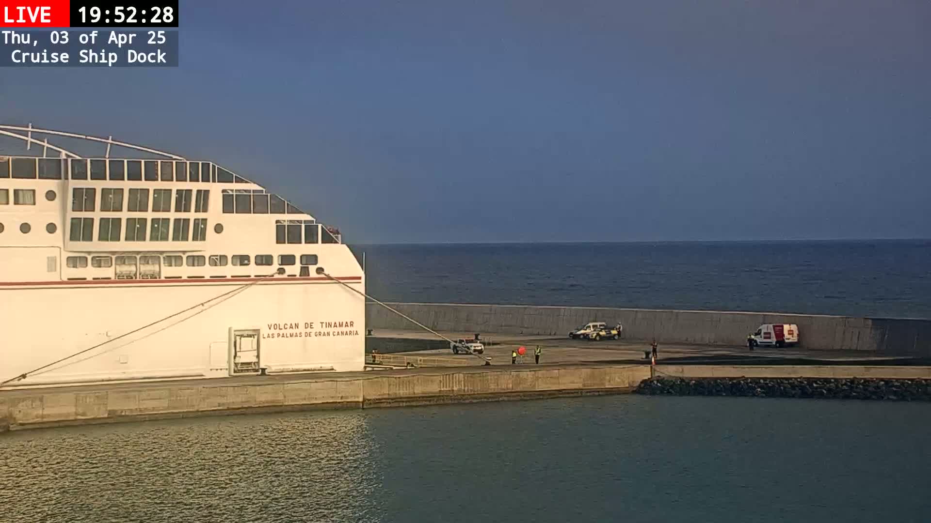 A cruise ship is docked at a port under a clear, twilight sky, with several vehicles and a few people visible on the dock.