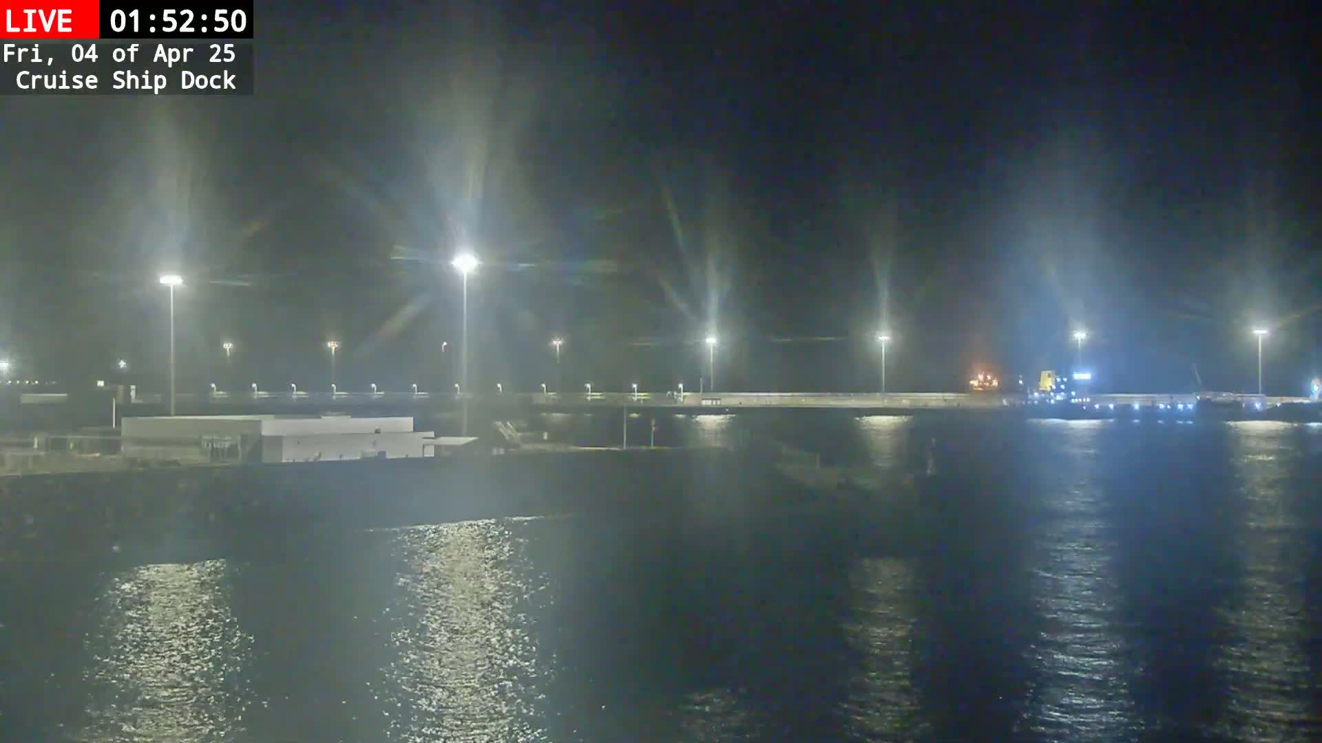 A cruise ship dock is illuminated at night by numerous bright lights, with a large vessel docked and calm water reflecting the lights.