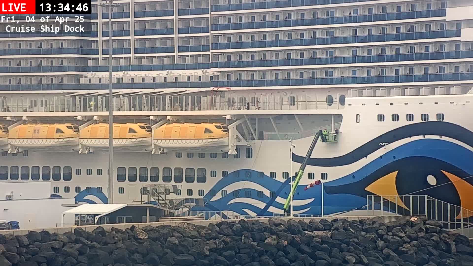 A large cruise ship with orange lifeboats is docked, with workers visible on a lift painting its hull; the weather appears to be sunny.