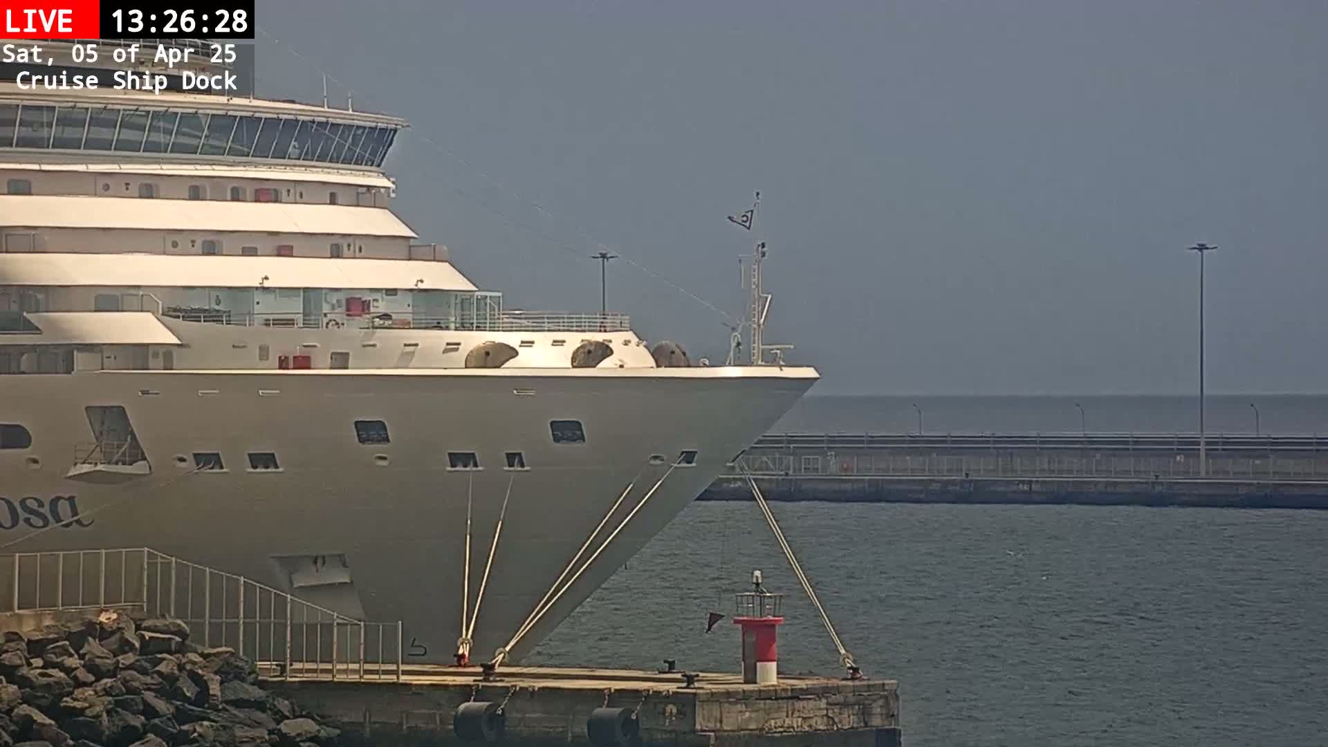 The bow of a large cruise ship is docked, secured by ropes to a pier on a calm, hazy day.