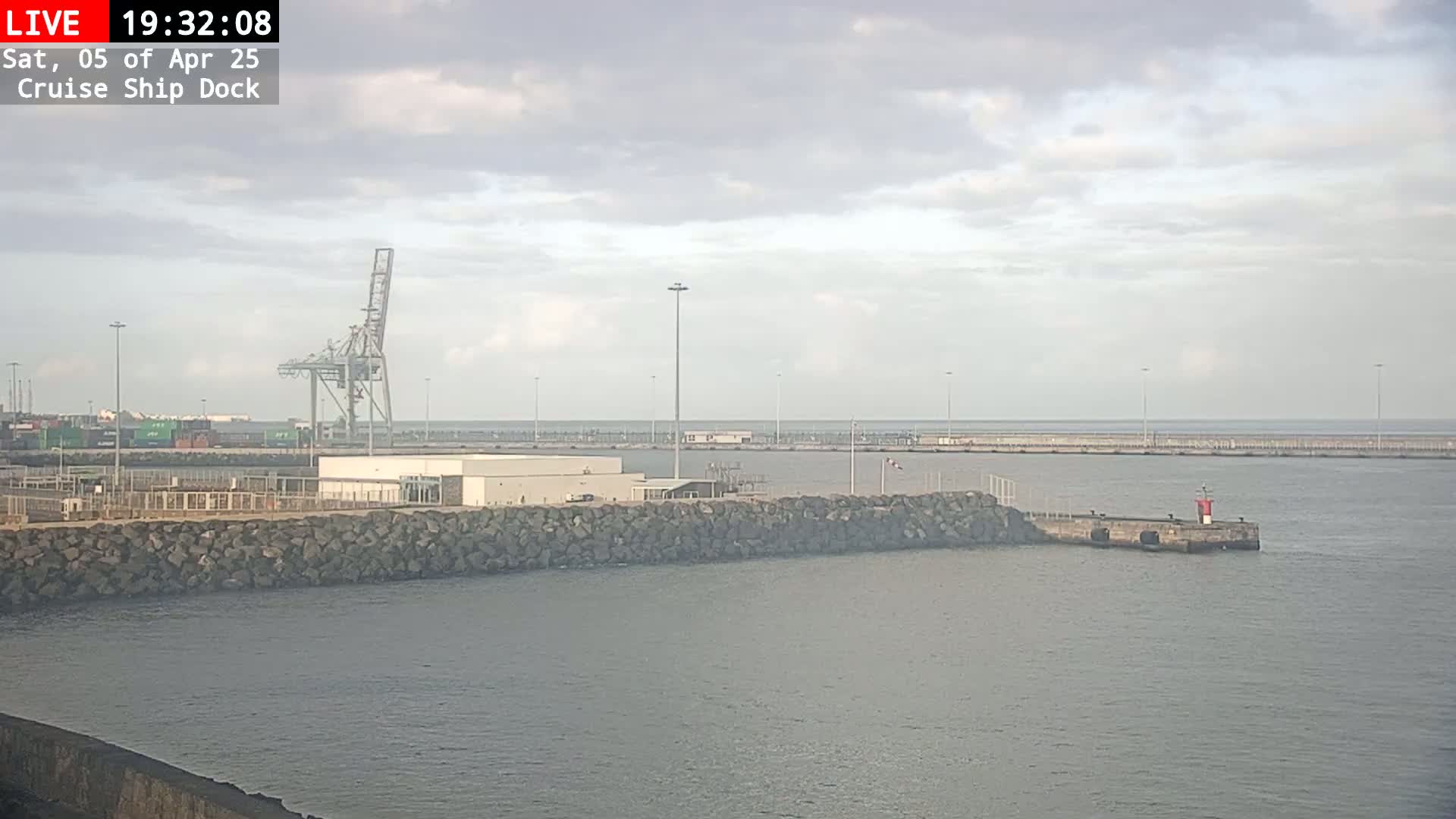 A cruise ship dock with a large crane and a rocky seawall is visible under a mostly cloudy sky.