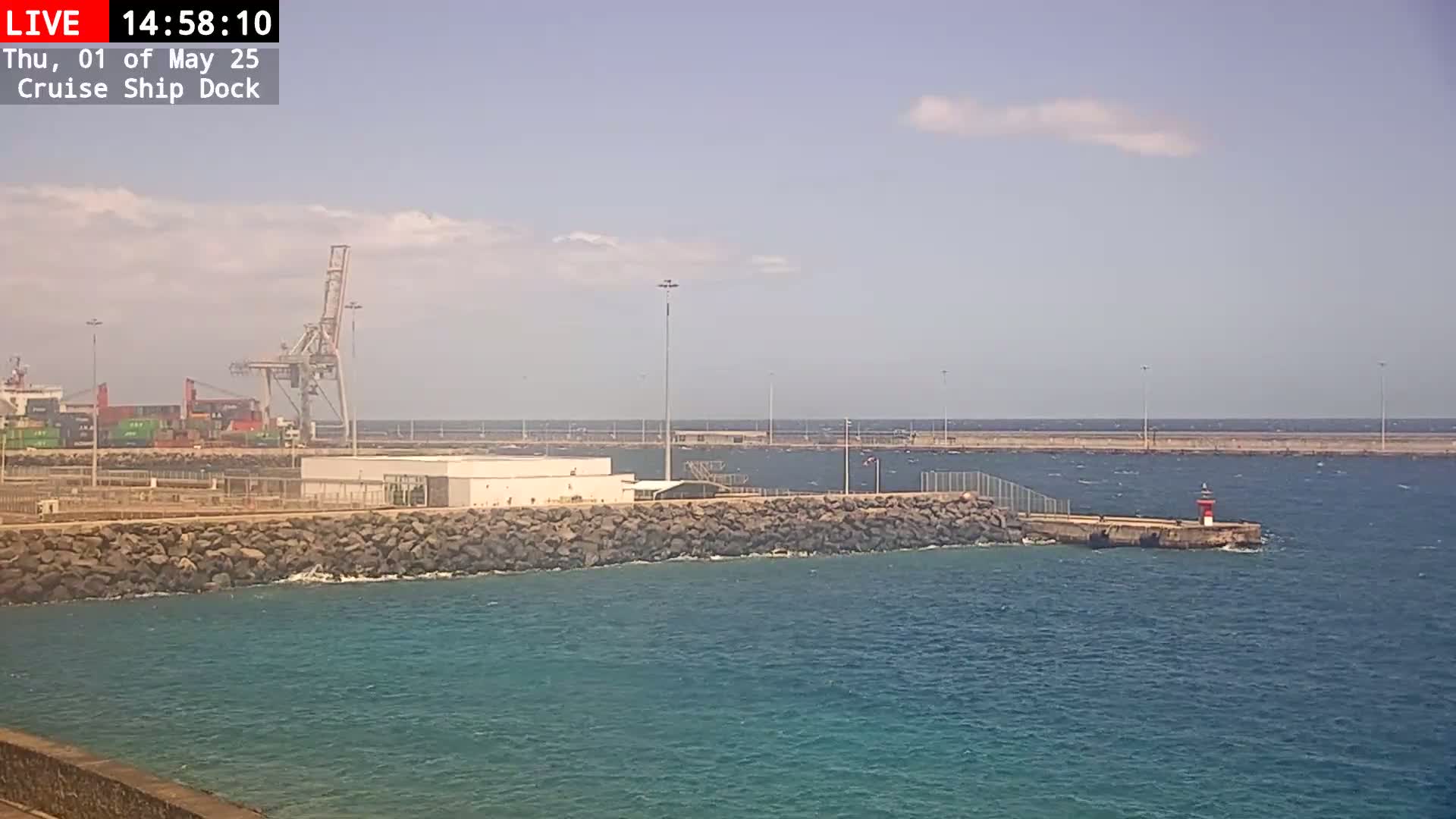 A partly sunny day shows a cruise ship dock with a cargo ship, cranes, and a small lighthouse in the water.