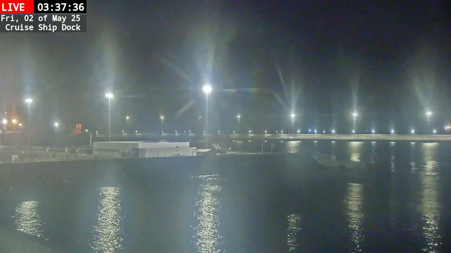 A nighttime view of a cruise ship dock, illuminated by many bright lights, reflecting on calm water.