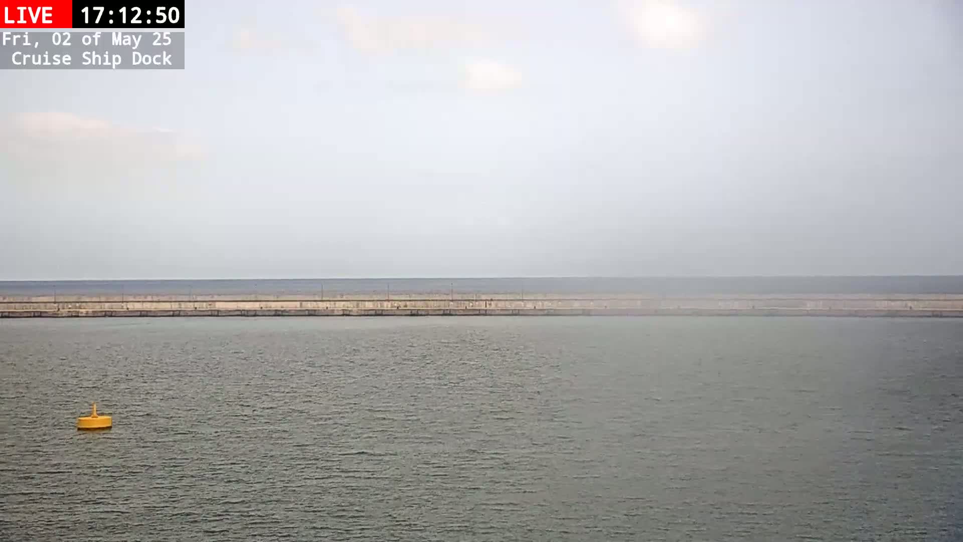 A calm, hazy body of water is seen beyond a long, low wall, with a single yellow buoy visible in the foreground.