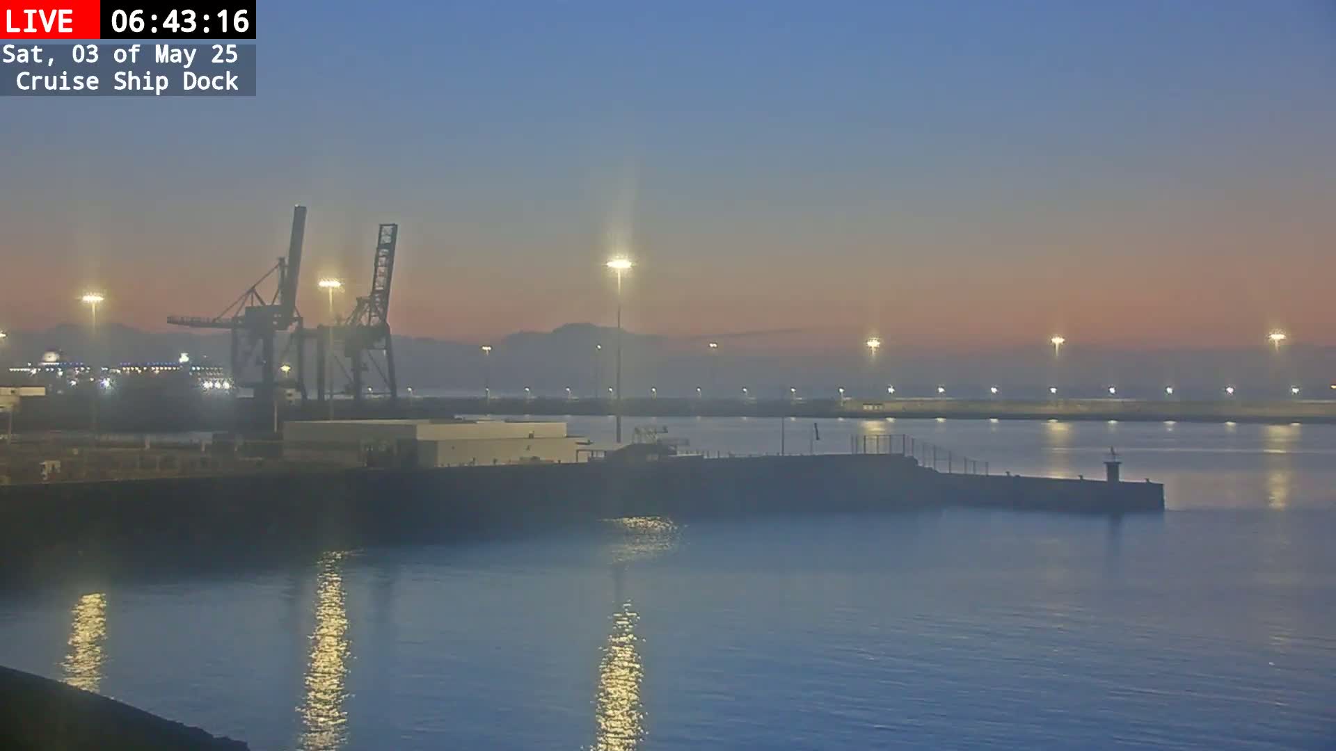A hazy sunrise illuminates a harbor with several cranes and lights reflecting on calm water.