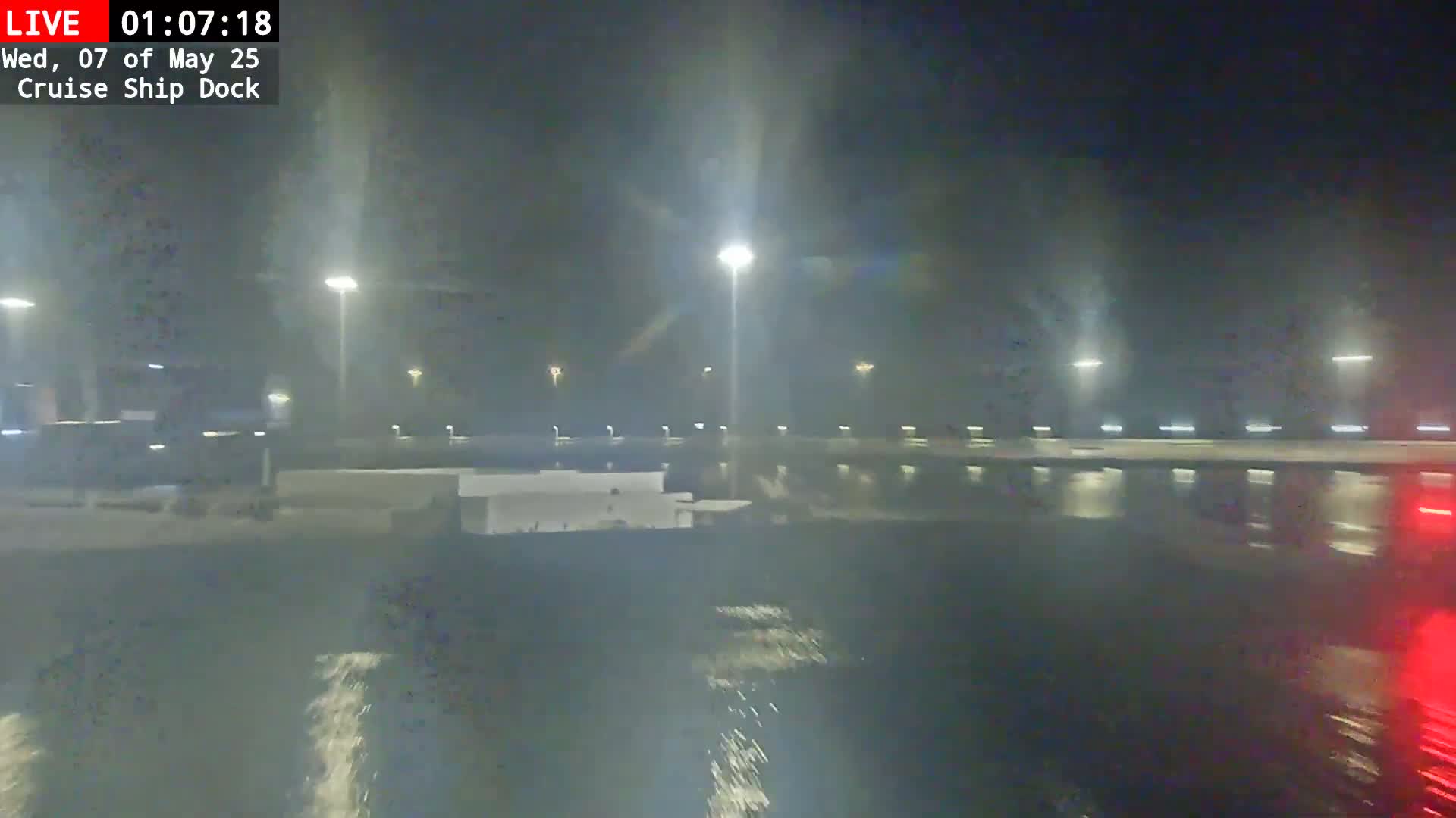 A nighttime view of a rain-soaked cruise ship dock with illuminated buildings and streetlights visible through the downpour.