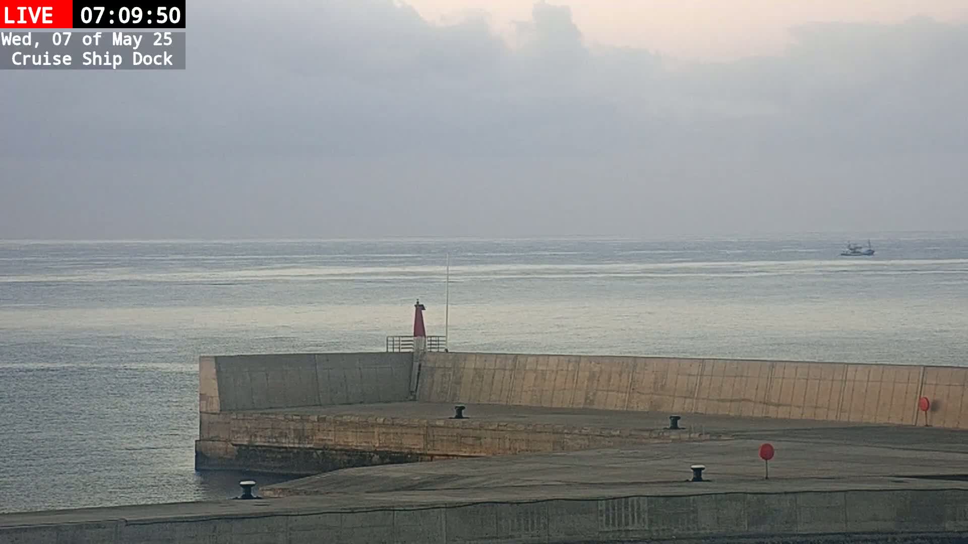 A concrete pier juts into a calm, gray sea under an overcast sky, with a small boat visible in the distance and a red lighthouse on the pier.