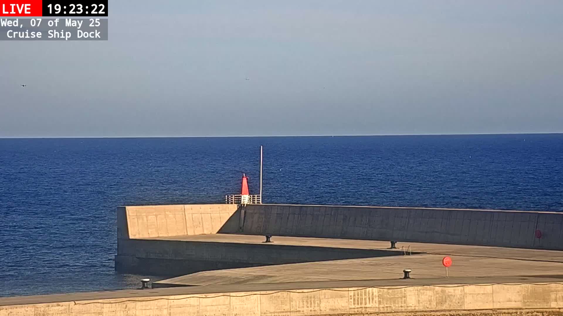 A concrete dock extends into a calm, blue sea under a clear sky, with a small red navigational marker visible.