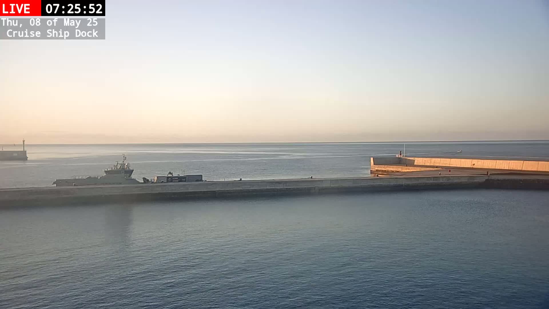 A calm, light-colored body of water shows a docked ship next to a long concrete pier under a pale sunrise.