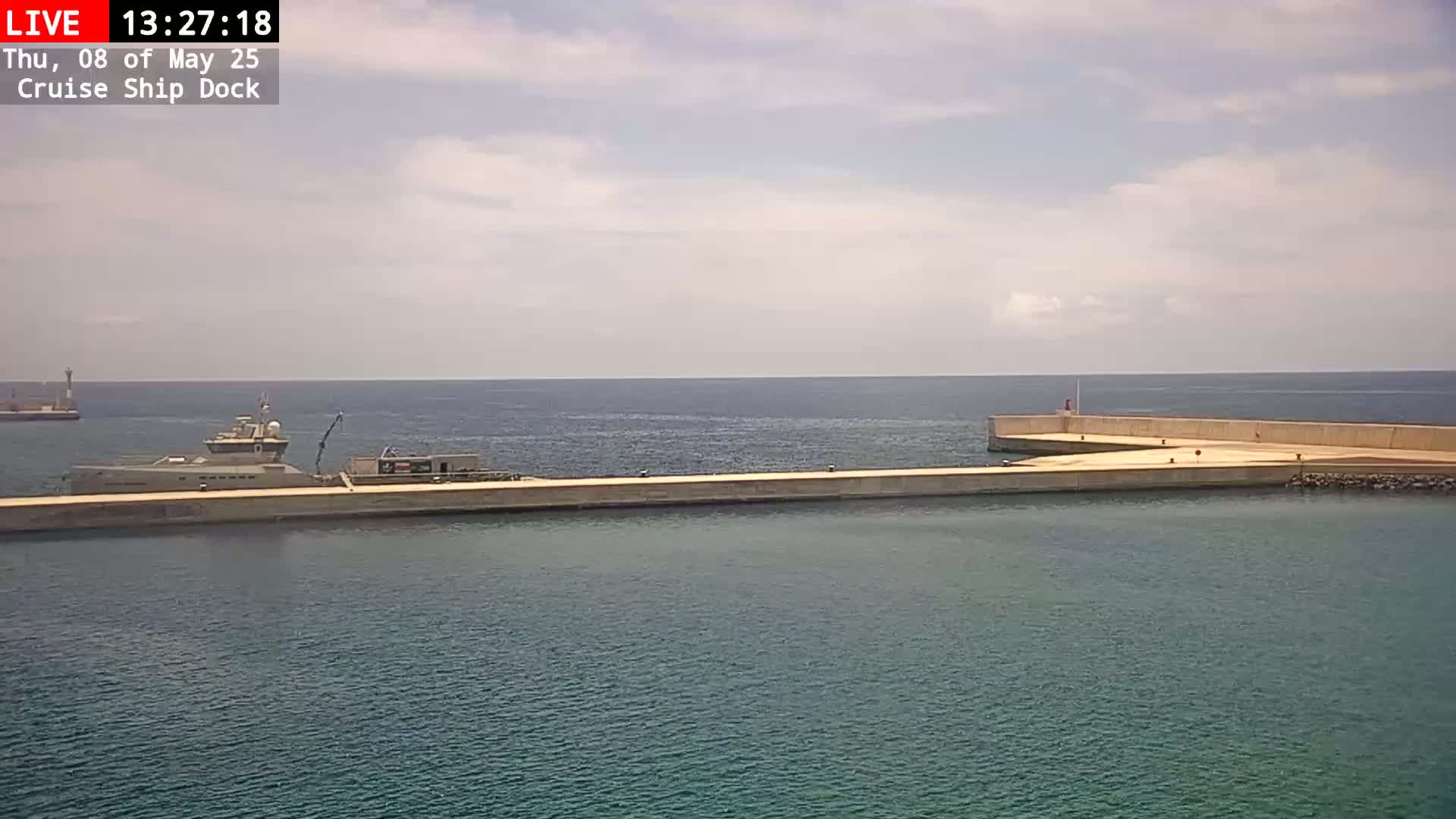 A large ship is docked at a concrete pier on a partly cloudy day, with calm water surrounding it.