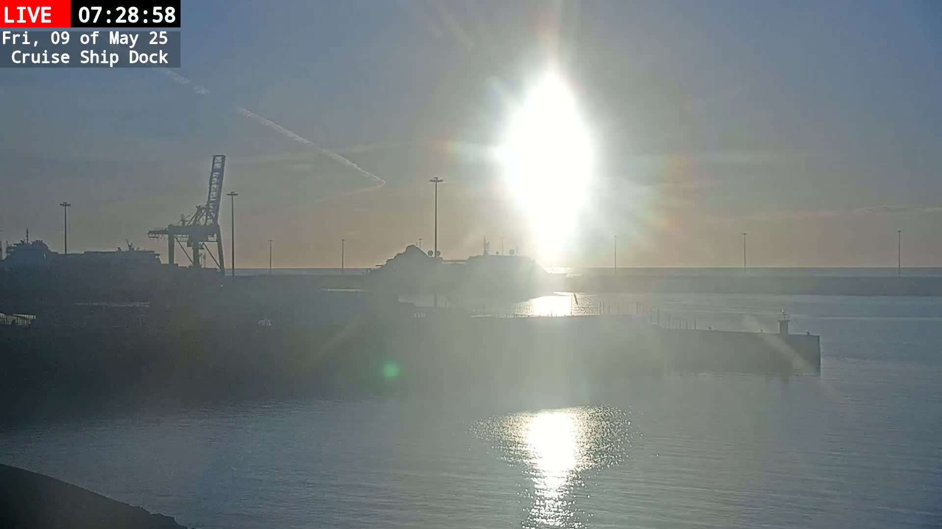 A brightly lit sunrise over a calm harbor with a docked cruise ship, a crane, and a breakwater.